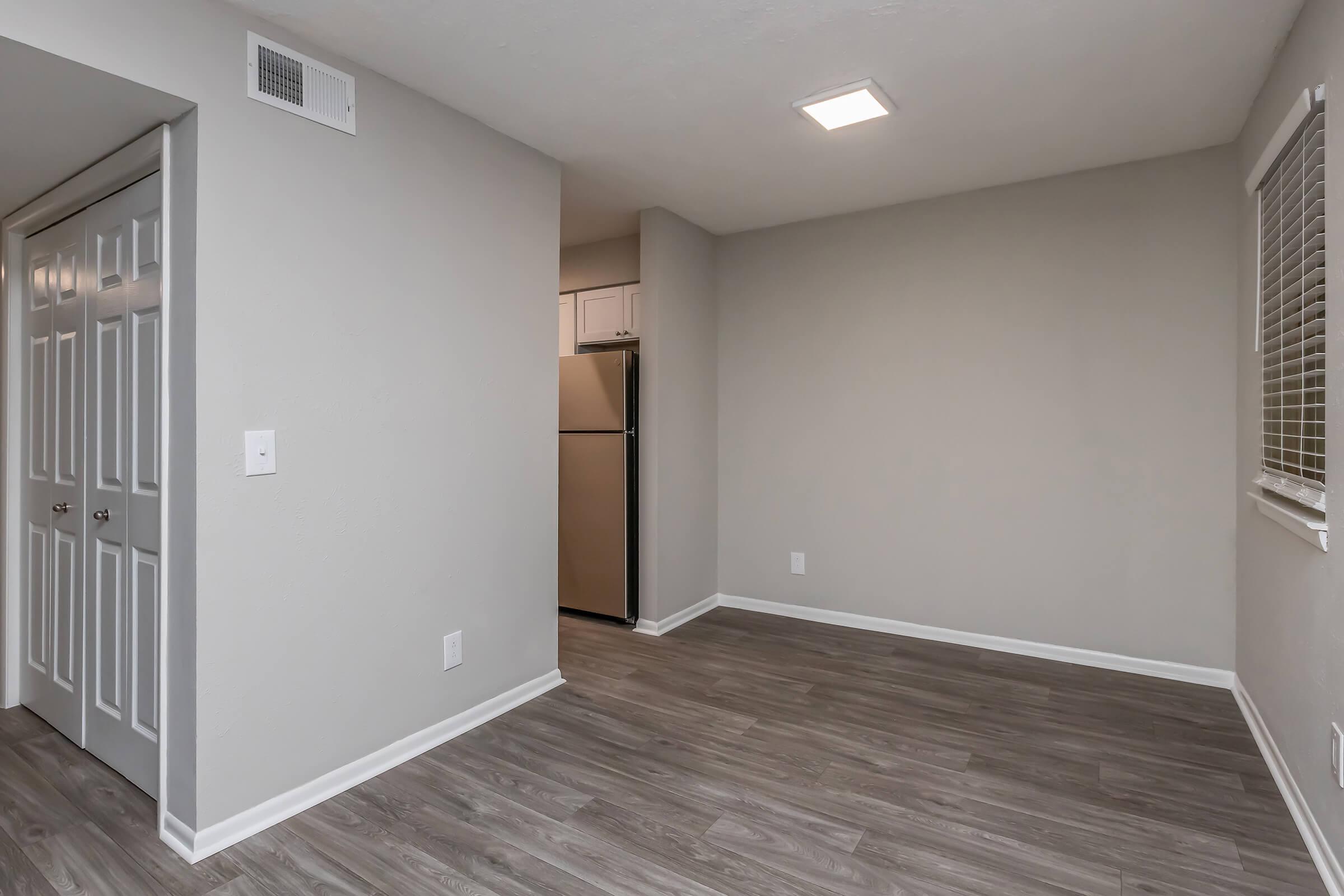 A spacious, empty room with light gray walls and laminate flooring. To the left, there are double doors leading to another space. In the background, a modern kitchen area is visible with a stainless steel refrigerator. Natural light comes in from a window on the right.