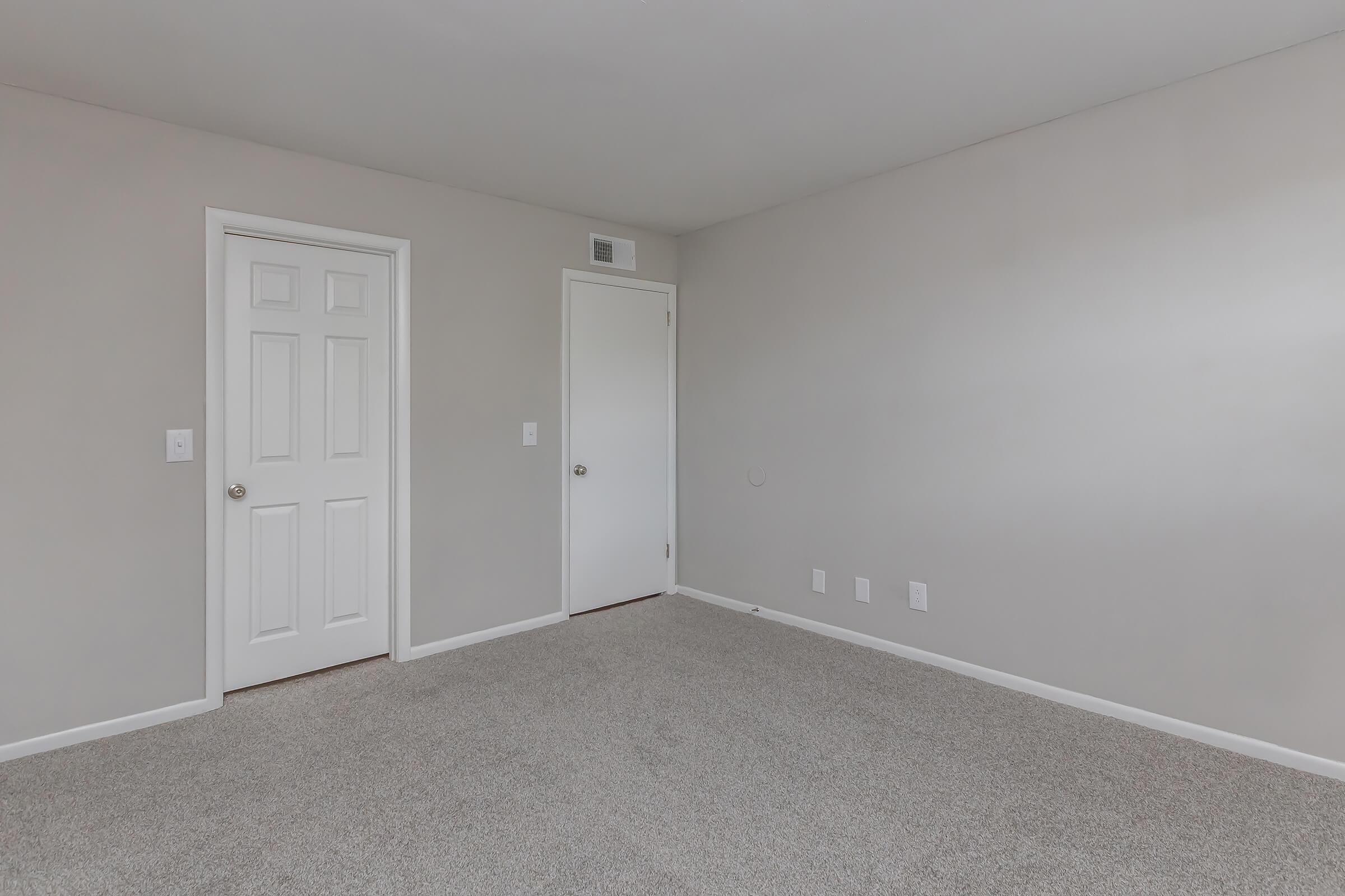 Empty room with light gray walls and a beige carpet. There are two closed doors: one white door on the left and another door on the right. The room has no furniture or decorations, creating a spacious and minimalistic appearance. Natural light may be coming from an unseen window.