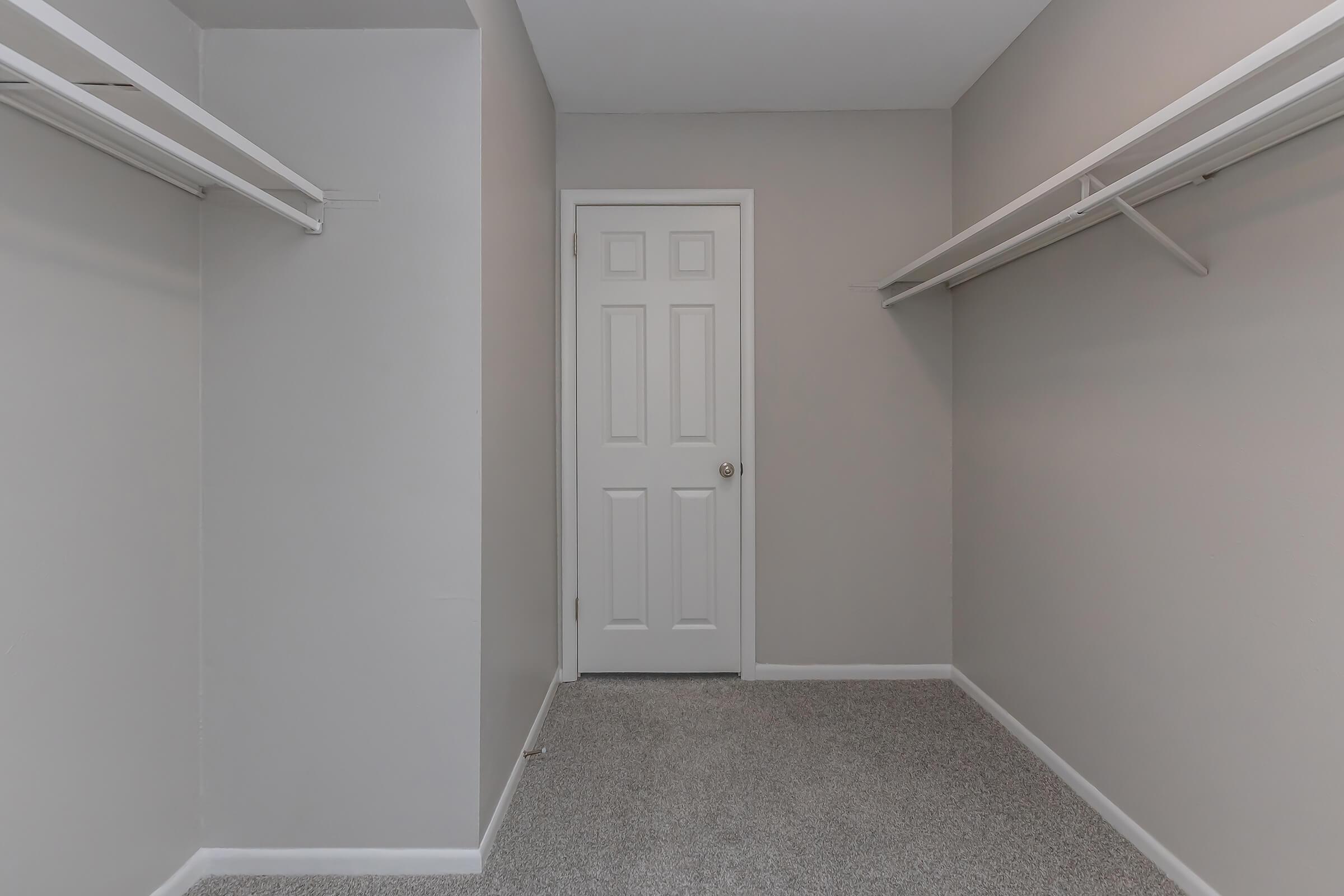A spacious, empty closet featuring light gray walls, two shelves on either side, and a closed white door at the end. The floor is covered in grey carpet, and the overall atmosphere is clean and minimalist.