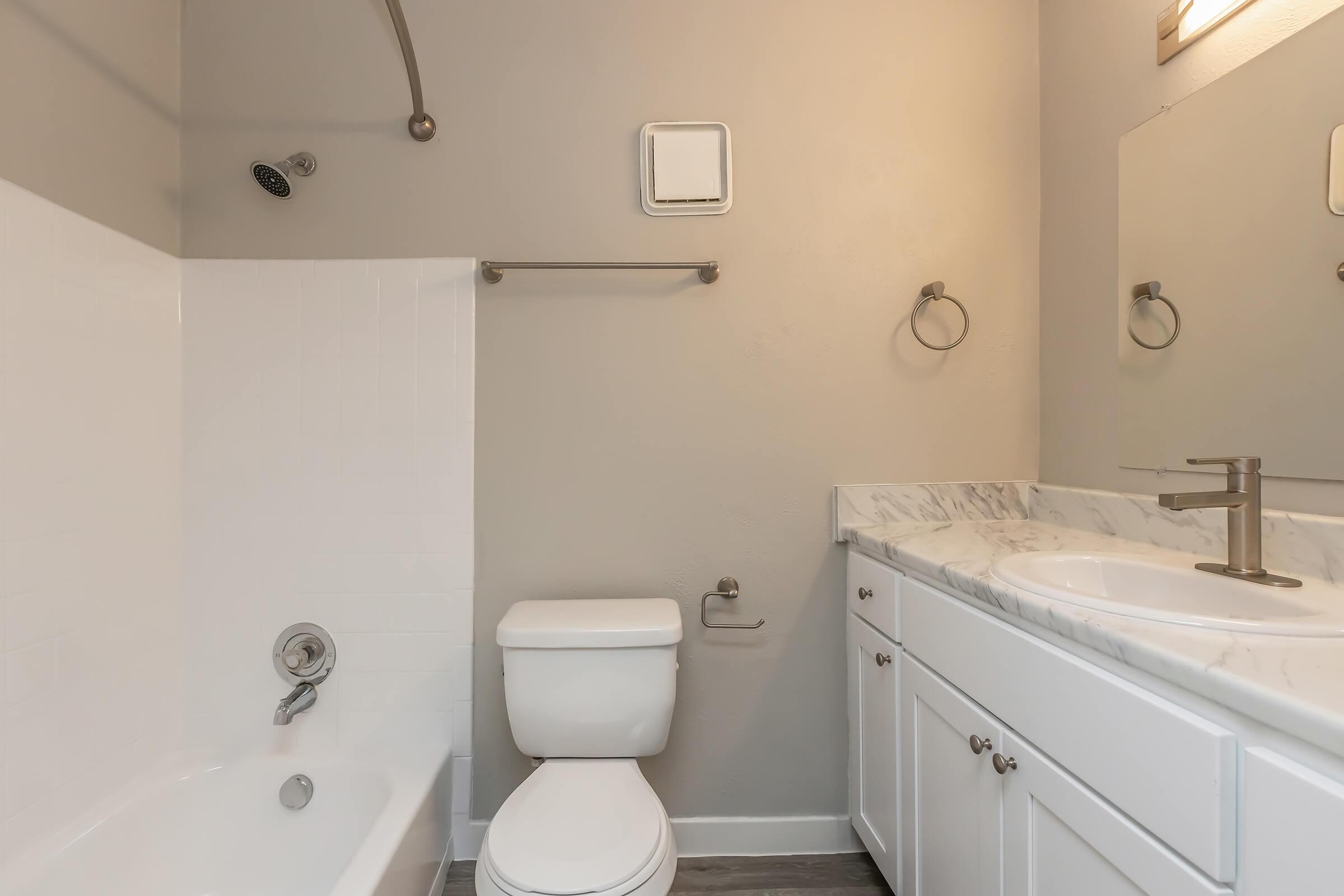A modern bathroom featuring a bathtub with a shower, a toilet, and a white double vanity with a marble countertop. Light gray walls complement the white fixtures. The space includes towel bars and a large mirror above the sink, creating a clean and contemporary look.