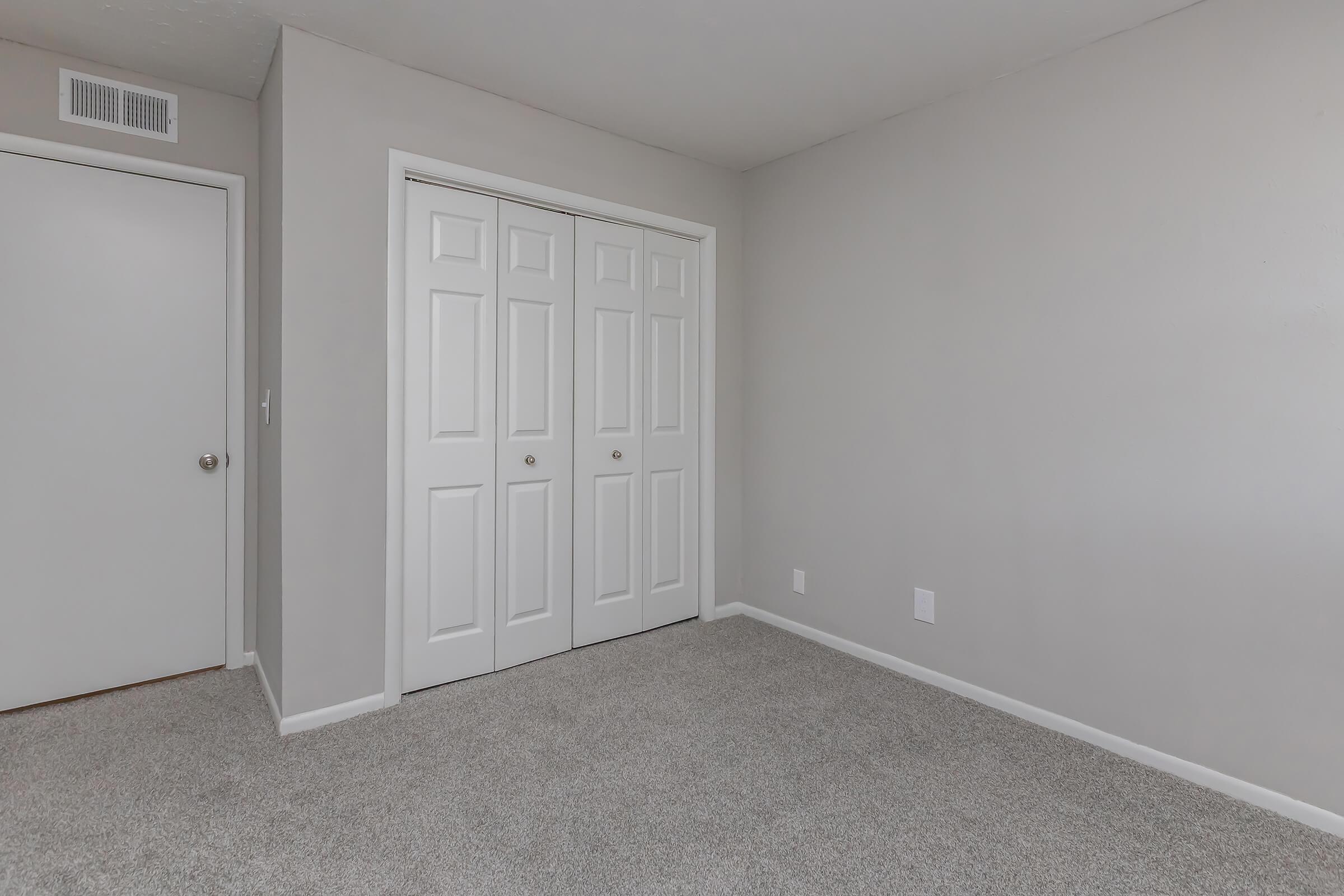 A simple, empty room with light gray walls and carpet. It features a closet with double white doors and a closed door leading to another space. The room has minimal decor and natural light, creating a clean and spacious appearance.