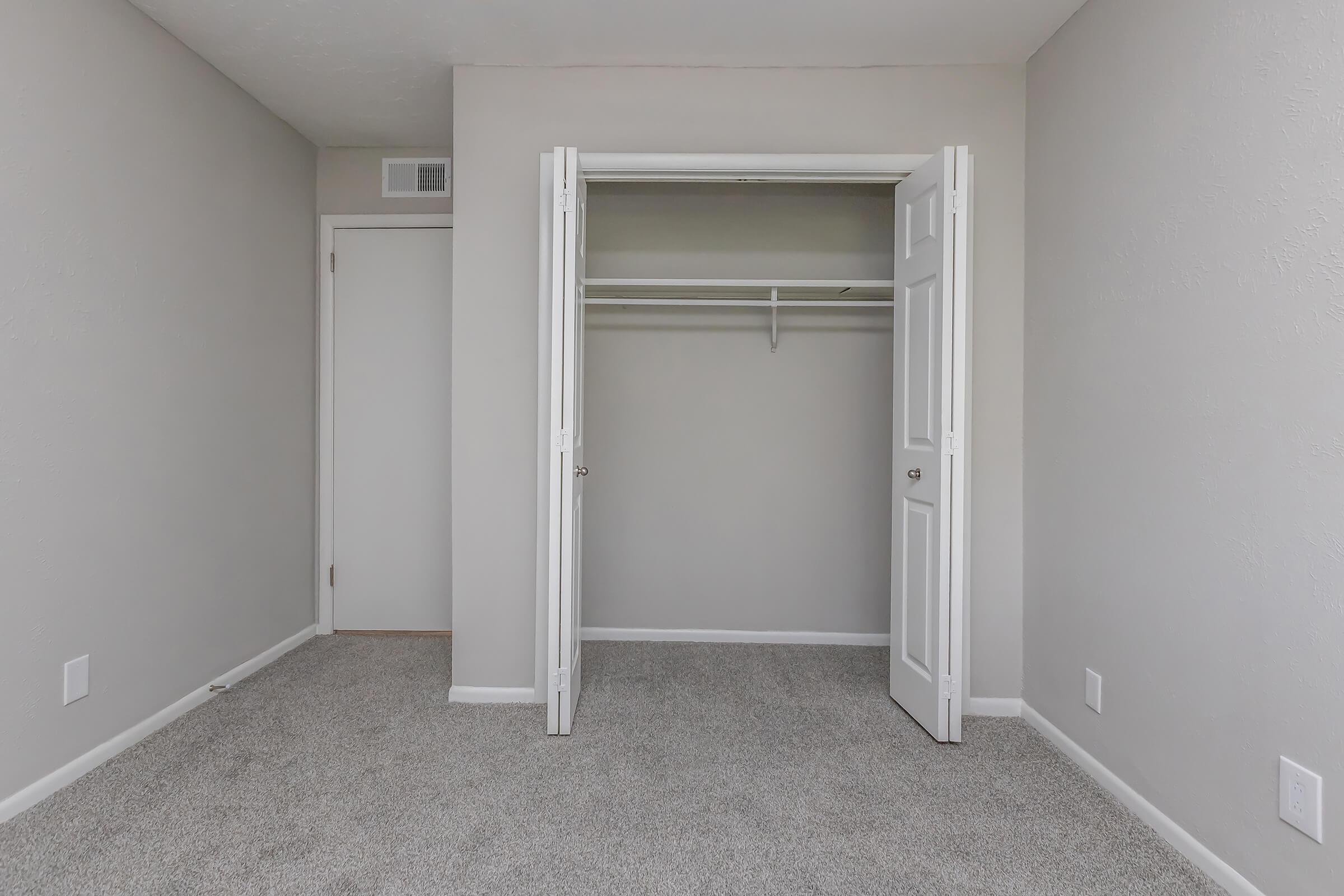A vacant room featuring light gray walls and a carpeted floor. There is an open closet with white double doors, revealing a single shelf inside. A closed door is visible on the left side, and the overall space appears clean and minimally furnished.