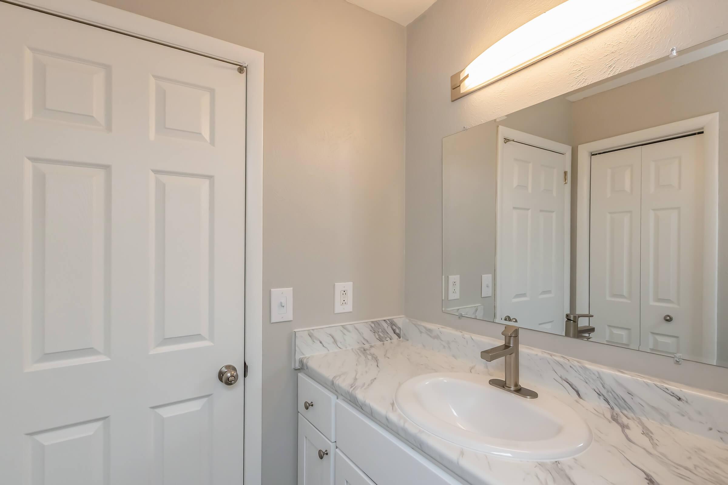 A modern bathroom featuring a white marble countertop with a circular sink, a contemporary faucet, a large mirror above the sink, and light gray walls. A closed white door is visible on the left, reflecting the mirror and the bathroom's clean design.