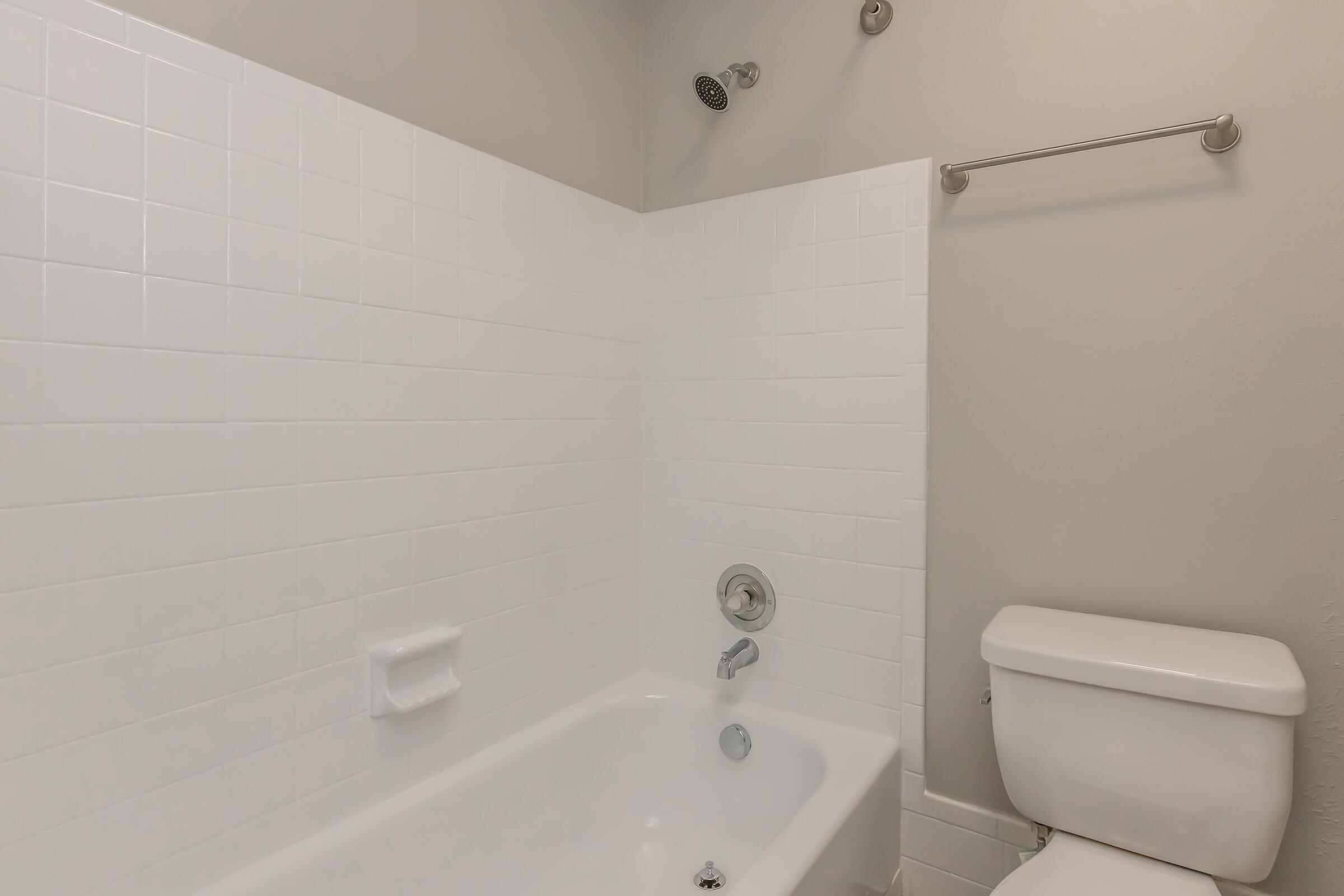 A clean bathroom featuring a white tiled shower area with a tub, a showerhead, and a chrome faucet. On the right, there is a white toilet, and above, a towel rack mounted on the wall. The walls are painted a light color, creating a fresh and modern atmosphere.