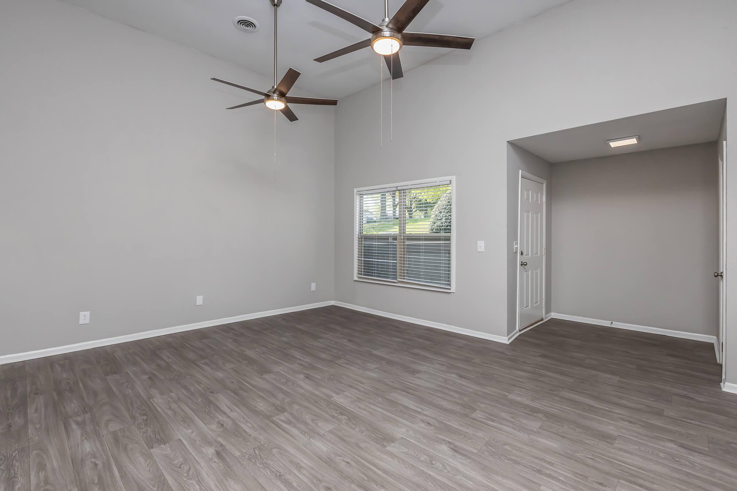 Spacious, empty interior of a room with light-colored walls and wooden flooring. Two ceiling fans are mounted above. A large window allows natural light to enter, and there’s an open door leading to another room. The overall atmosphere is bright and airy.