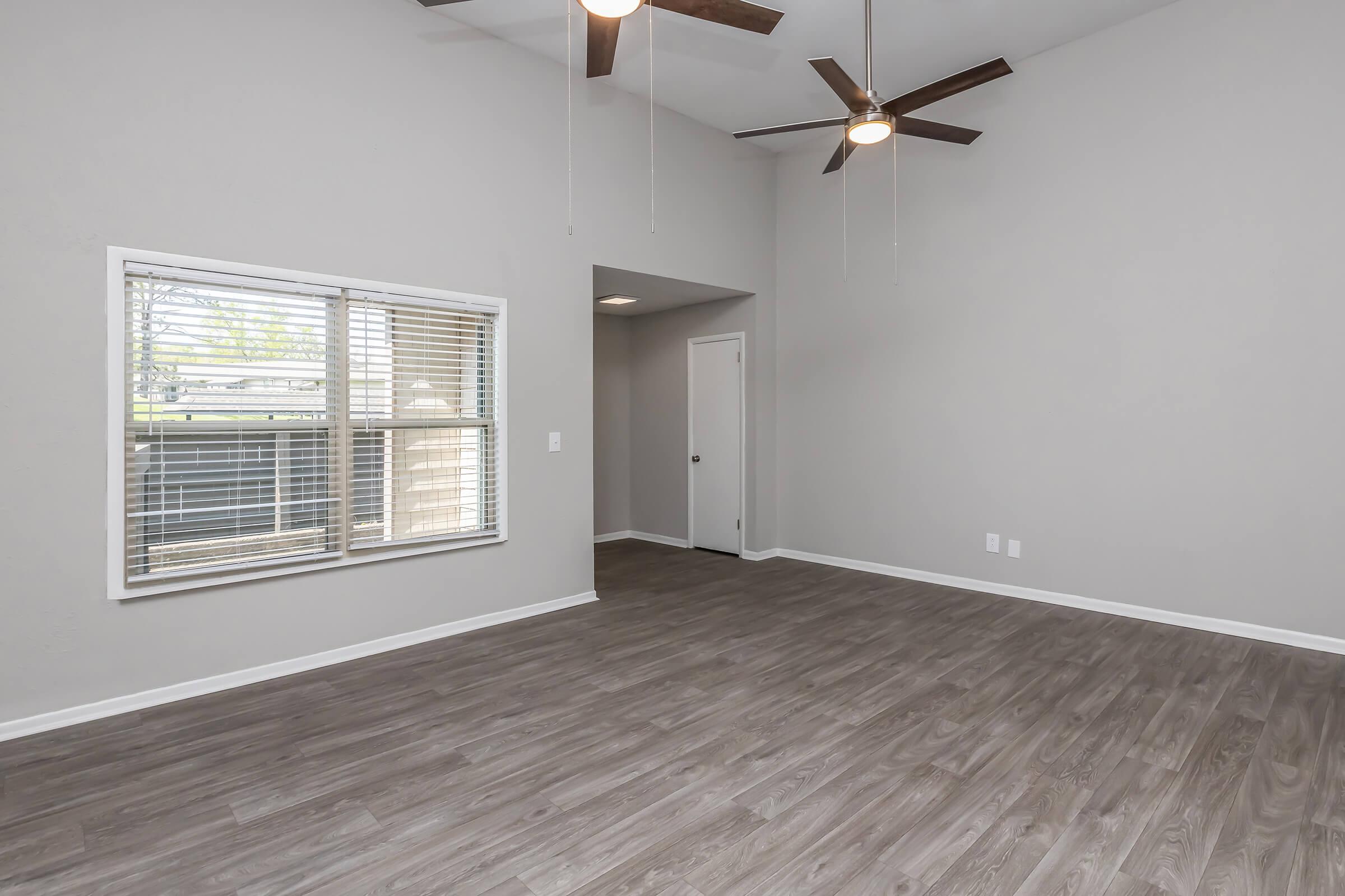 Spacious interior of a modern living room featuring light gray walls, vinyl flooring, and two ceiling fans. A large window on one wall allows natural light in, while a door leads to another room. The room is empty, highlighting its openness and potential for various layouts.