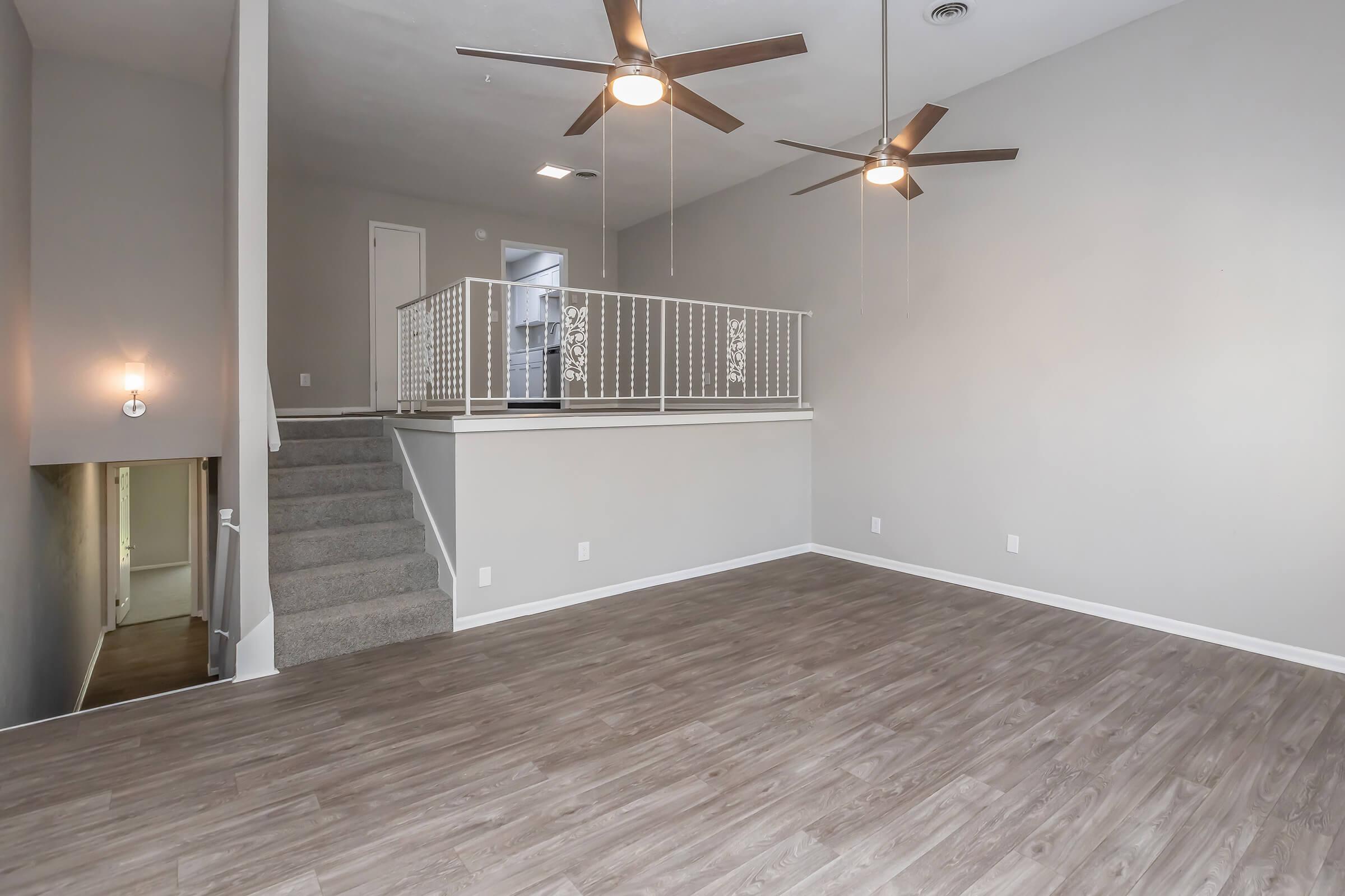 Spacious living area featuring light wood flooring, two ceiling fans, and a staircase leading to a loft-style balcony. The walls are painted in a soft gray tone, enhancing the open feel of the space. A stairwell leads down to a lower level, adding additional living space.