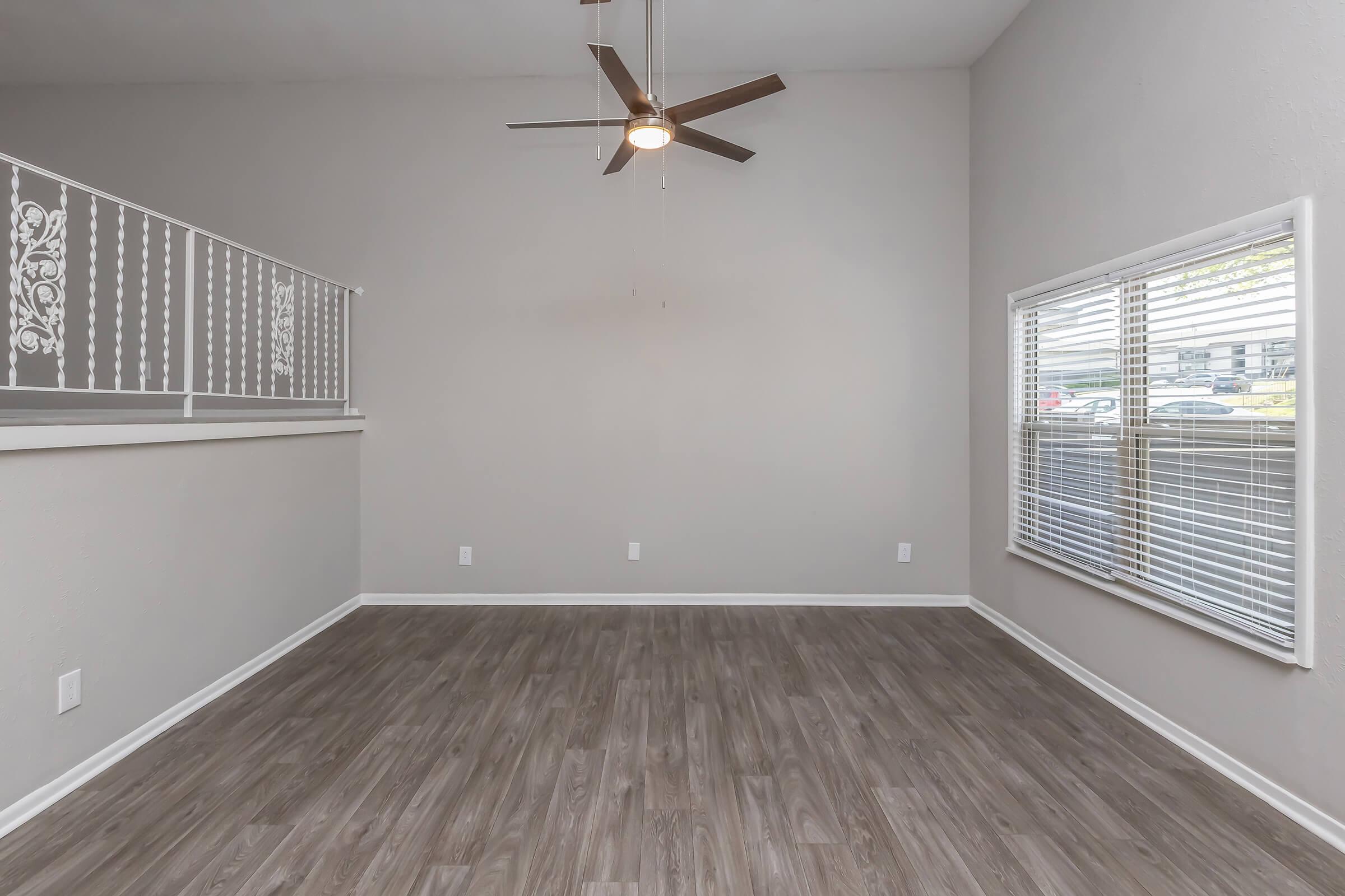 Spacious living area with grey walls and wood-like flooring. A ceiling fan hangs above, and large windows with blinds allow natural light. A stair railing is visible, leading to an upper level. The room is empty, creating a blank canvas for potential furnishings and decor.