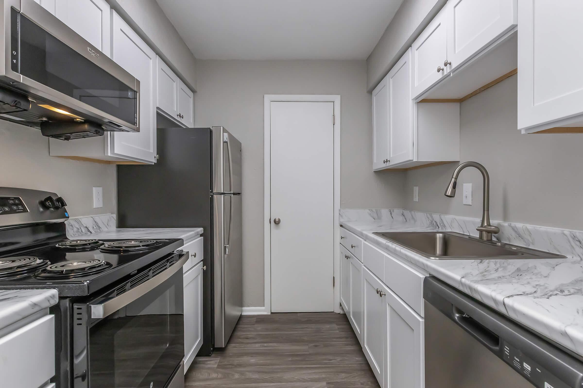 A modern kitchen featuring stainless steel appliances, including an oven, microwave, and refrigerator. The countertops are made of marble, and there are white cabinets above. A stainless steel sink is integrated into the countertop, with a door visible leading to another area of the home.