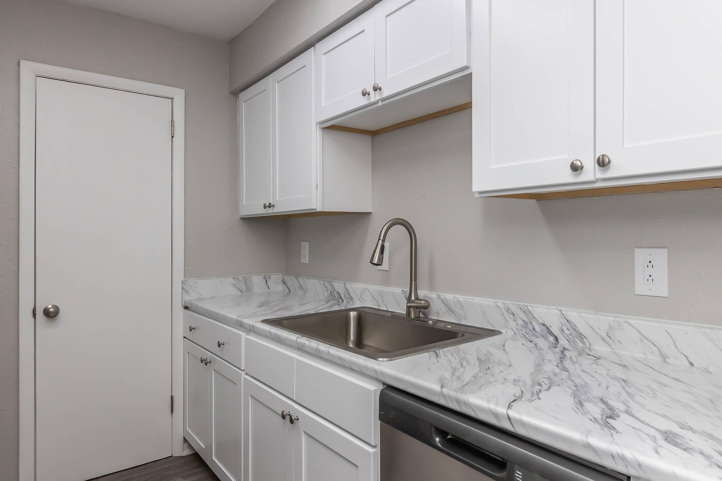 A modern kitchen with white cabinetry, a marble countertop, and a stainless steel sink. The kitchen features a dishwasher and a closed door leading to another room, all set against a neutral wall color. Bright and tidy, the space showcases contemporary design elements.