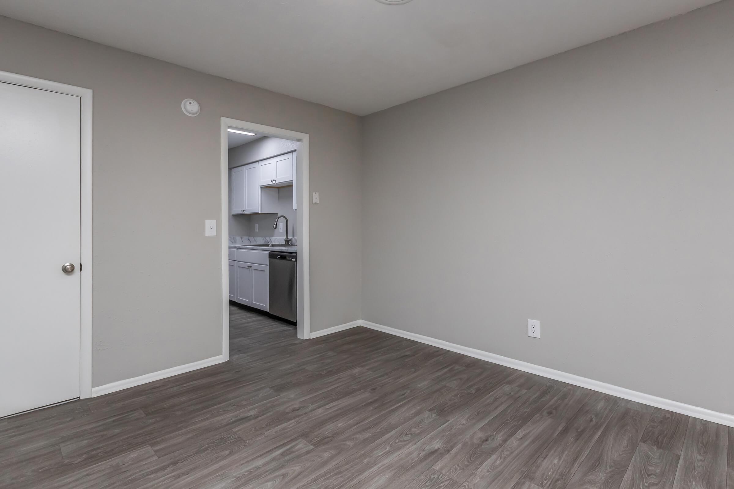 Interior view of a neutral-colored room with light gray walls and a wooden floor. A doorway leads to a kitchen area with white cabinets and appliances visible in the background. The space is empty, providing a sense of openness and potential for furnishing.