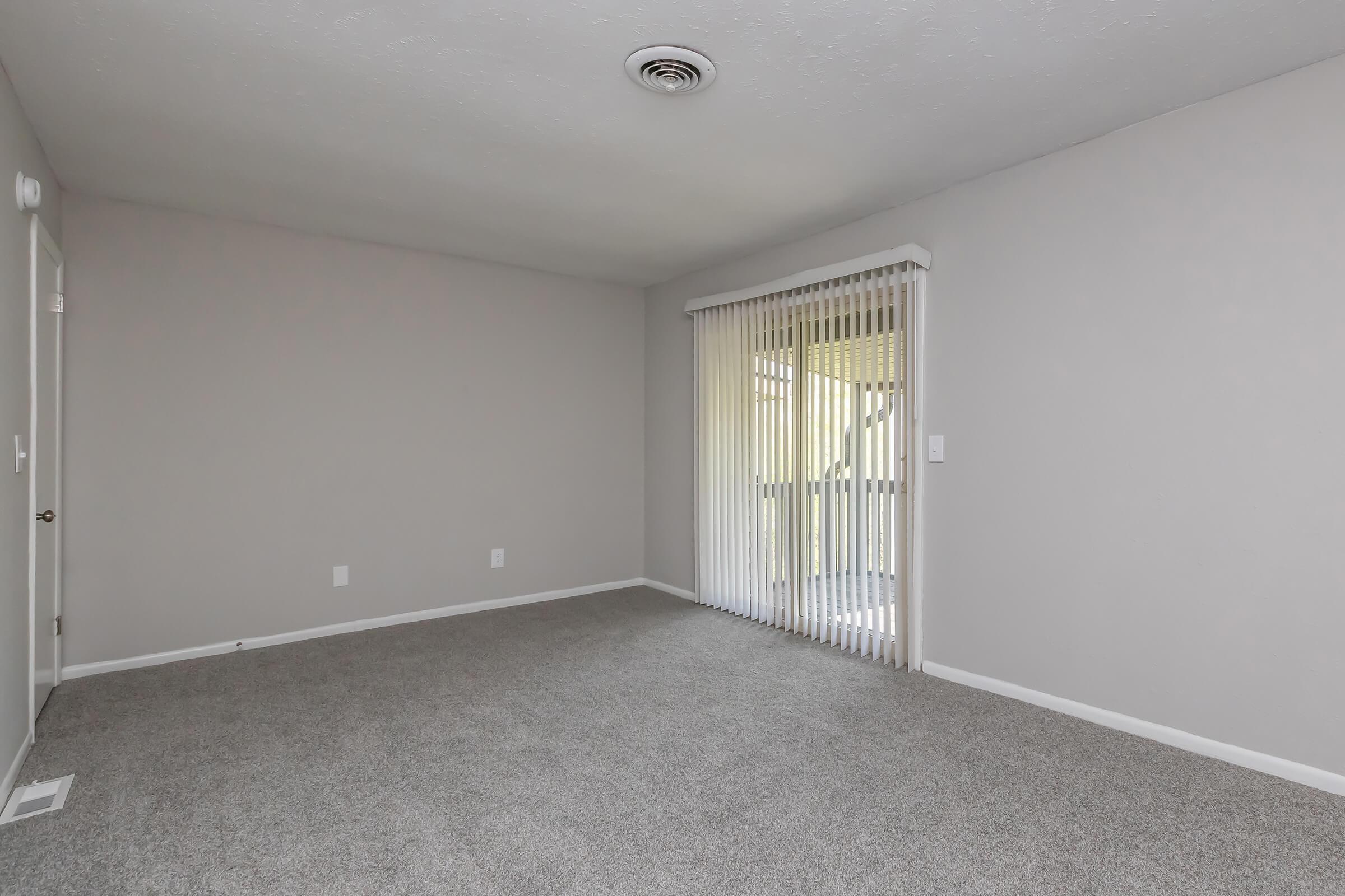 A spacious, empty living room featuring light gray walls, plush carpet, and large sliding glass doors with vertical blinds leading to an outside balcony. The room is well-lit, showcasing a neutral color palette with no furniture or decor. Ideal for personal customization.