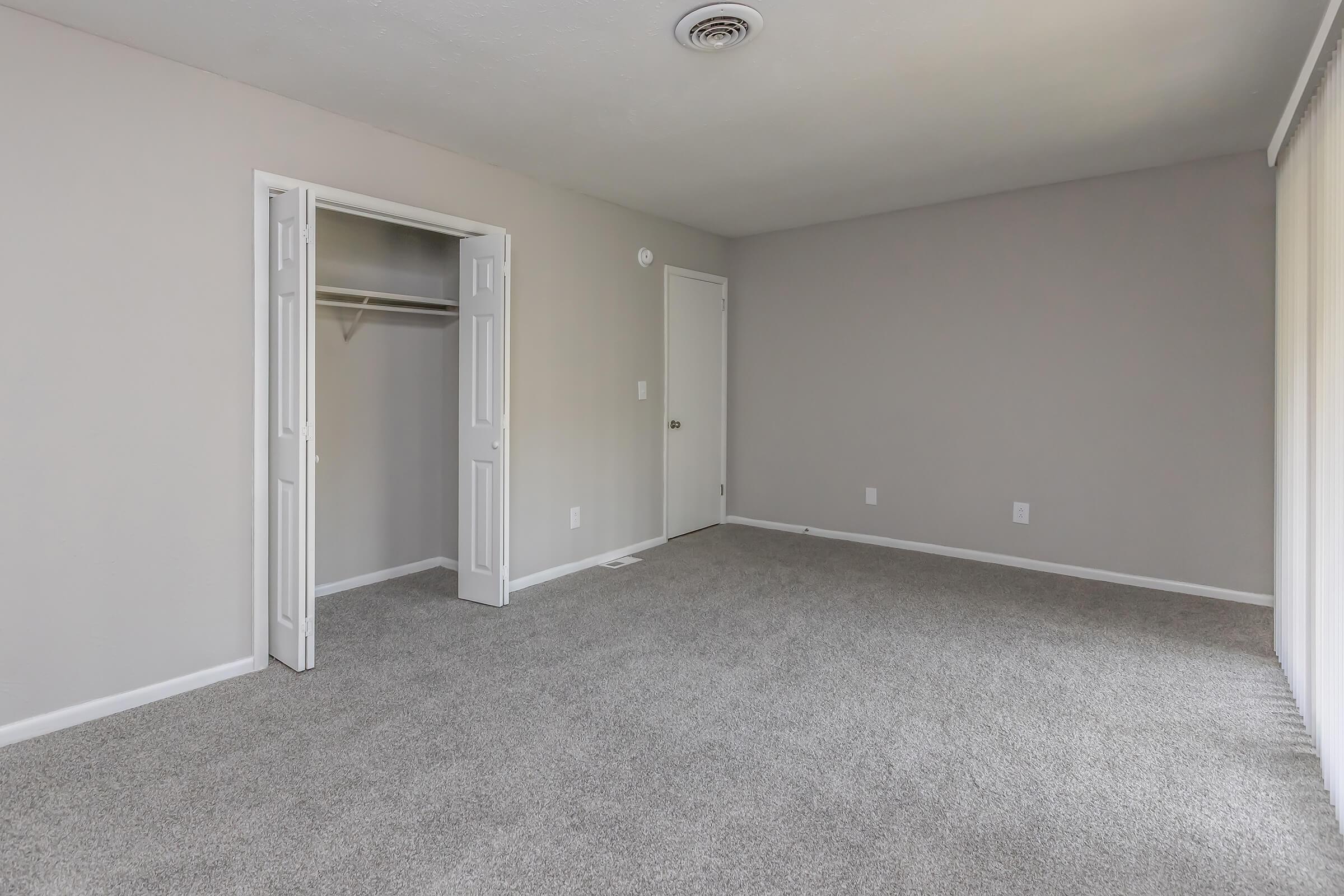 A spacious, empty room with light gray walls and carpet. There is a small closet with double doors on the left and a solid closed door on the right. Natural light enters through sliding glass doors on the opposite wall, which is partially covered by sheer curtains.