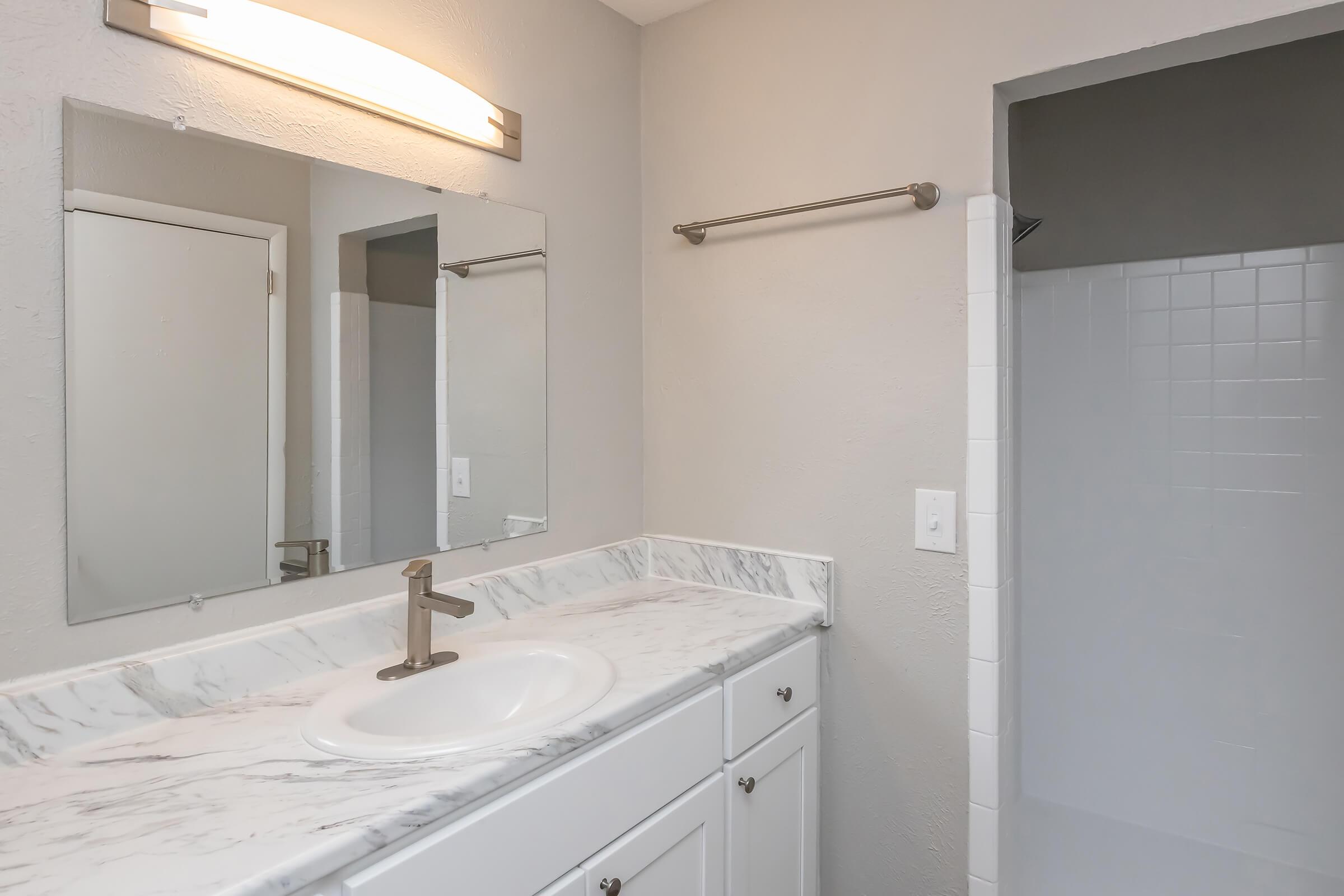 Modern bathroom featuring a marble countertop with an under-mount sink, a large mirror, and a light fixture above. White cabinetry and a chrome towel bar are present. A door leads to a shower area, with tiled walls and a clean, minimalistic design. The walls are painted in a soft, neutral color.