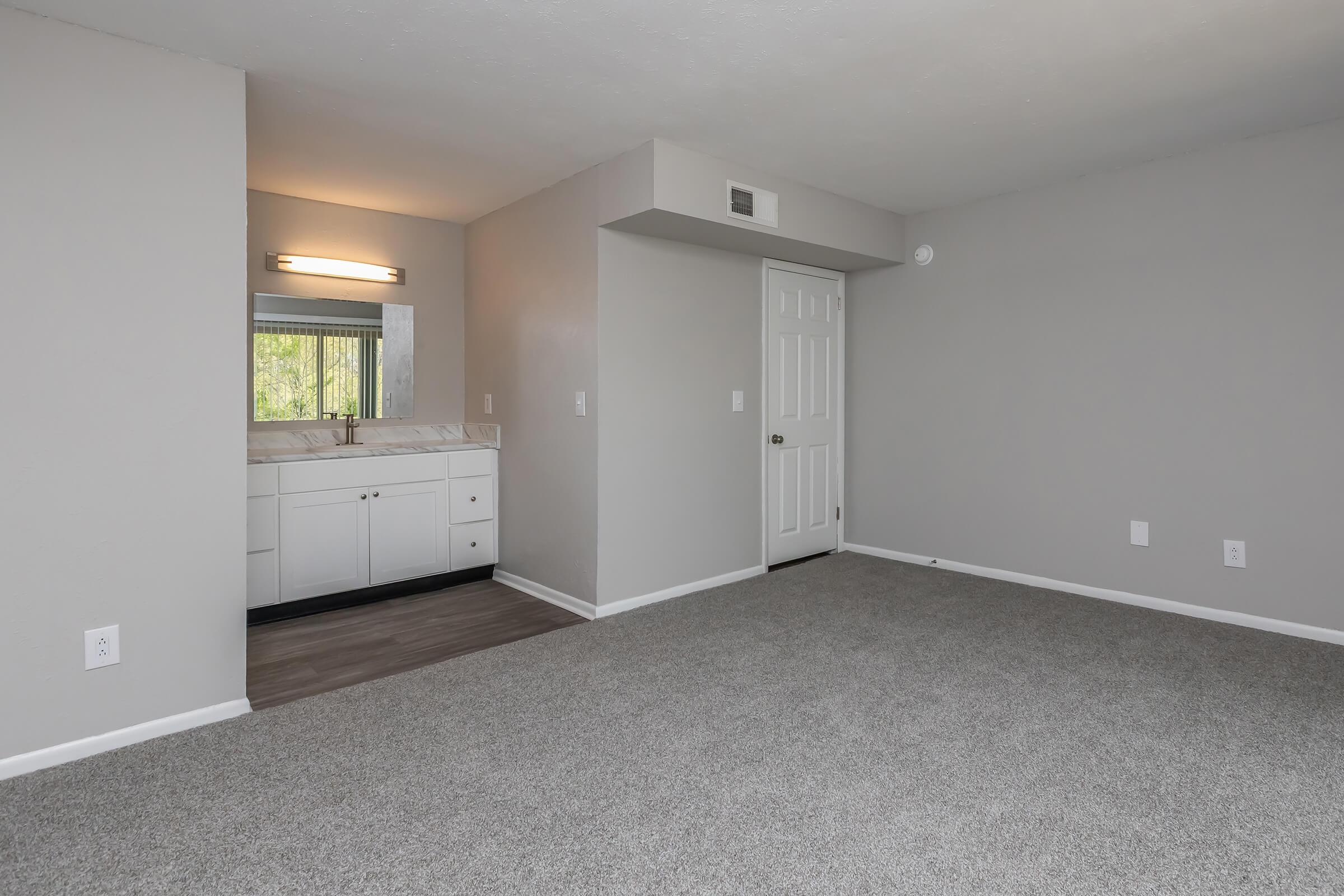 A spacious, clean room with light grey walls and carpet. On the left, there's a small area with a vanity and mirror, indicating a bathroom space. The room has a door leading to another area and a window with natural light, creating a bright and airy atmosphere.