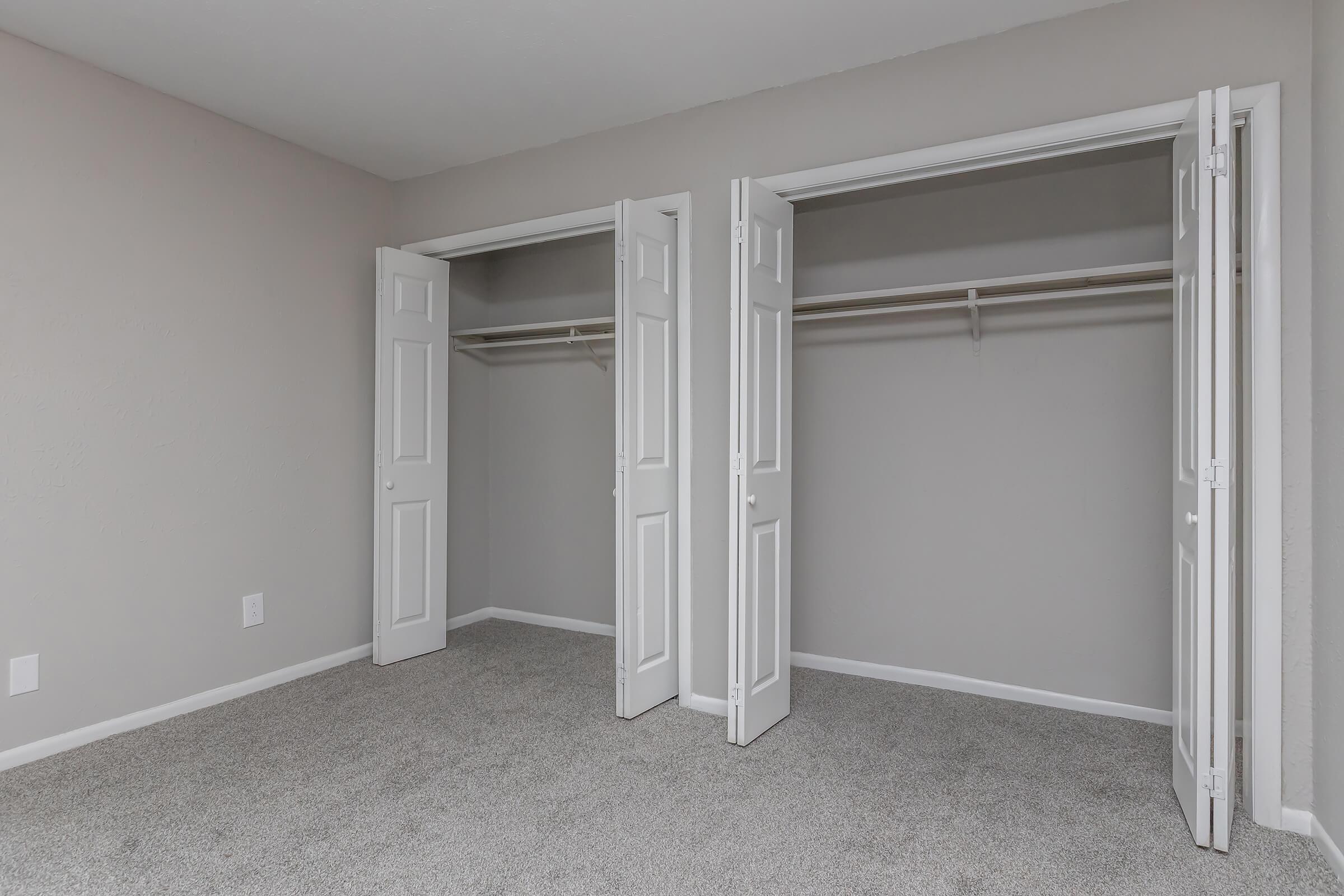 A light-colored room with gray walls and a carpeted floor. Two open closets with white doors are on the left and right, showcasing the empty shelving inside. The overall space is bright and minimalistic, emphasizing the closets as a functional storage area.