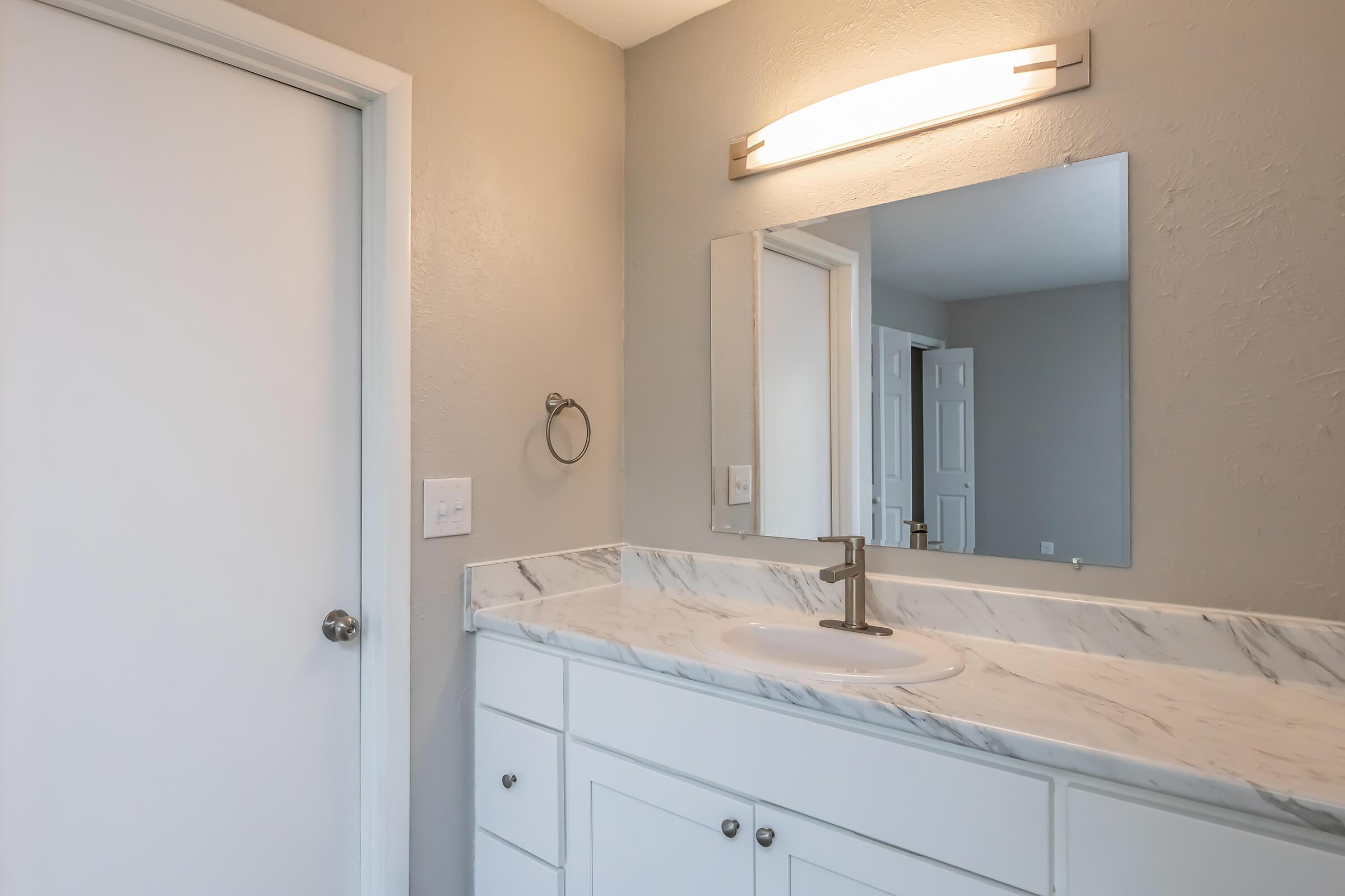 Bright bathroom with a simple design featuring a white marble countertop, a round sink with a modern faucet, a wall-mounted mirror, and a light fixture above. Light gray walls add to the clean aesthetic, with a door leading to another room visible in the background.