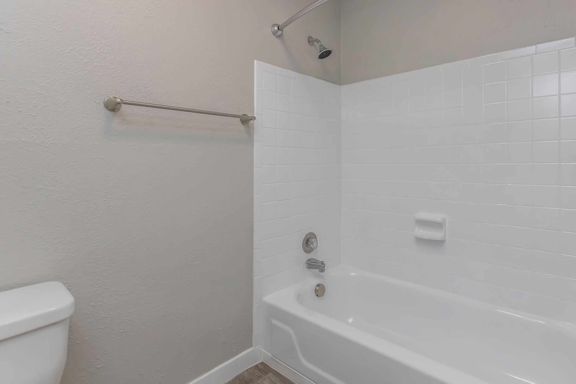 A clean, modern bathroom featuring a white bathtub with a showerhead, a tiled wall, and a silver towel bar. A white toilet is visible to the left, and neutral-colored walls create a minimalist aesthetic. The floor has a light wood-like appearance.