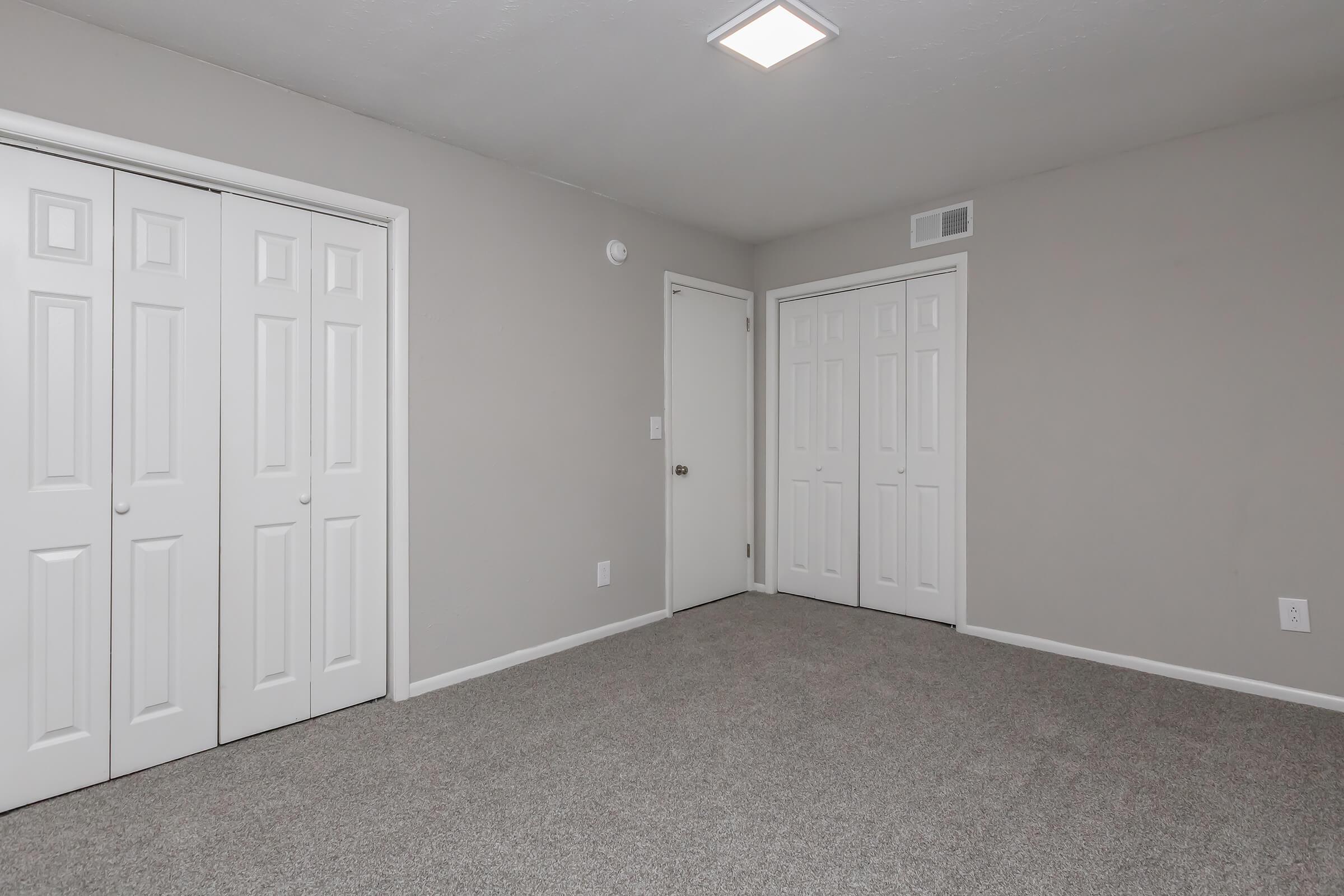 A spacious empty room with light gray walls and beige carpet. It features two sets of white double doors along one wall, a single white door on another wall, and a ceiling light fixture. The overall atmosphere is bright and minimalistic, suitable for various uses.