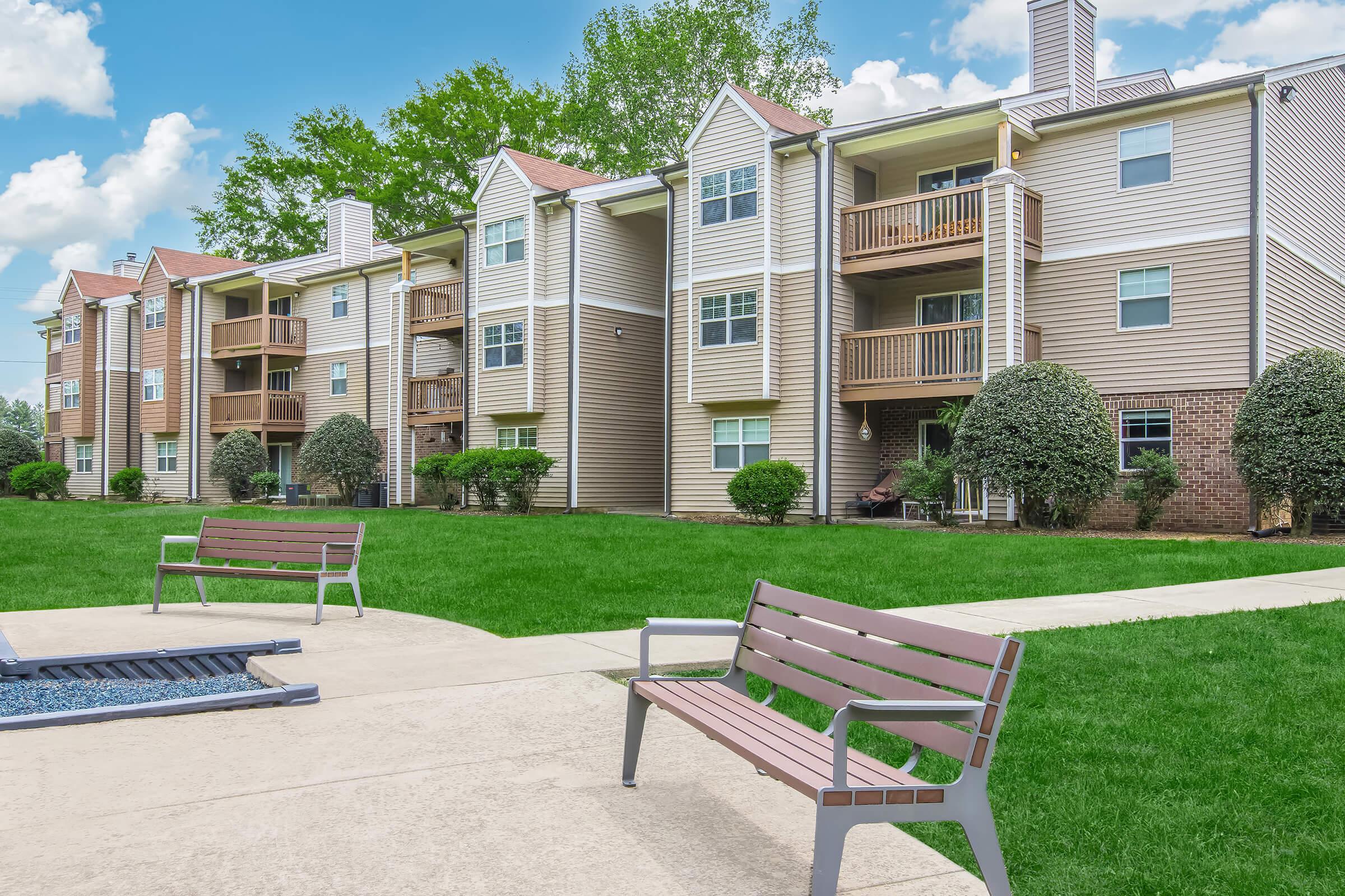 A residential apartment complex with multiple buildings featuring balconies. Well-maintained green lawns and landscaped shrubs surround the buildings. In the foreground, there are two benches facing a small patio area with a fire pit, set against a backdrop of a blue sky with scattered clouds.