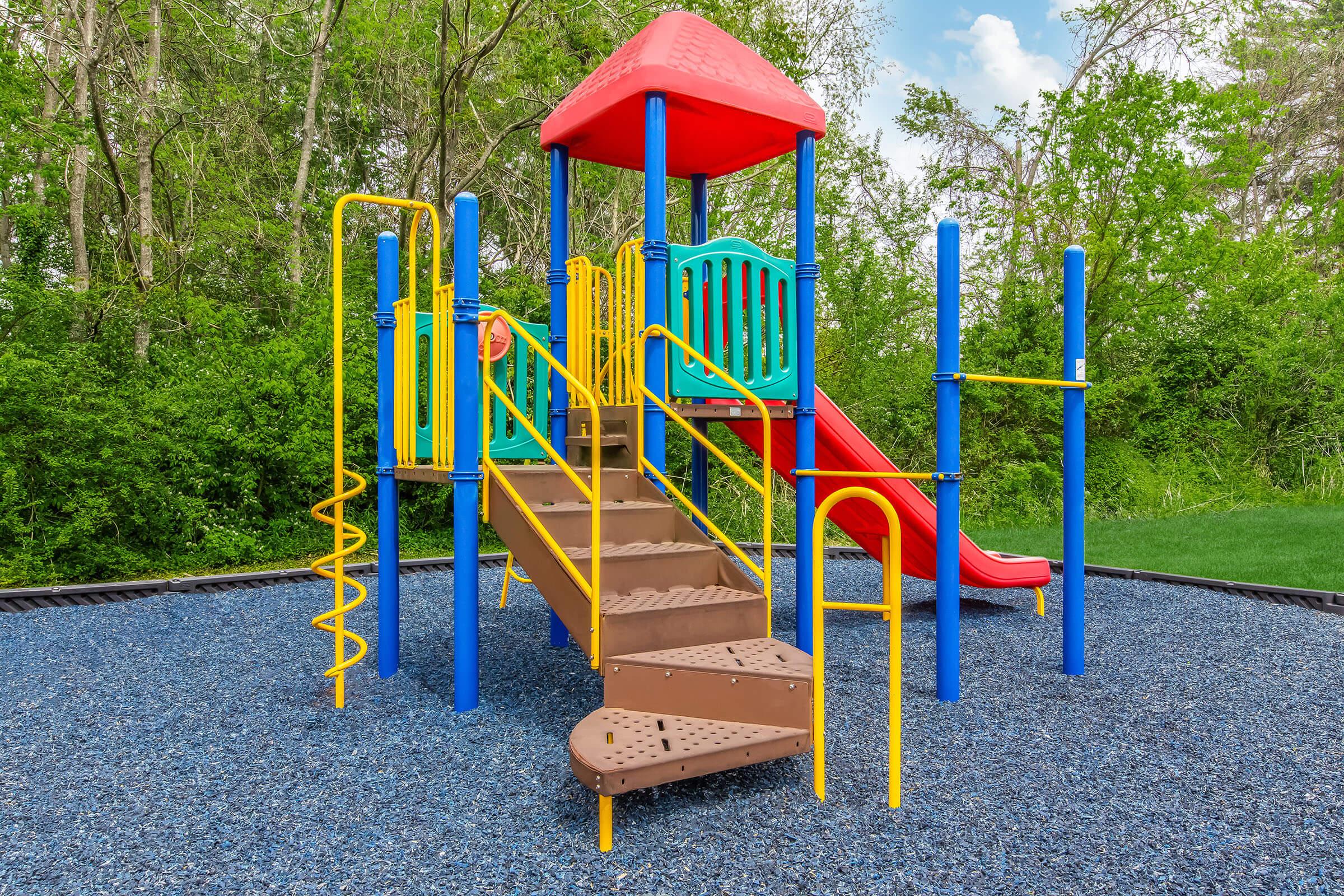 A colorful playground structure featuring a red and blue slide, yellow climbing bars, and stairs, set in a grassy area surrounded by trees. The play equipment is designed for children, with ample safety features and a soft ground covering made of blue rubber mulch.