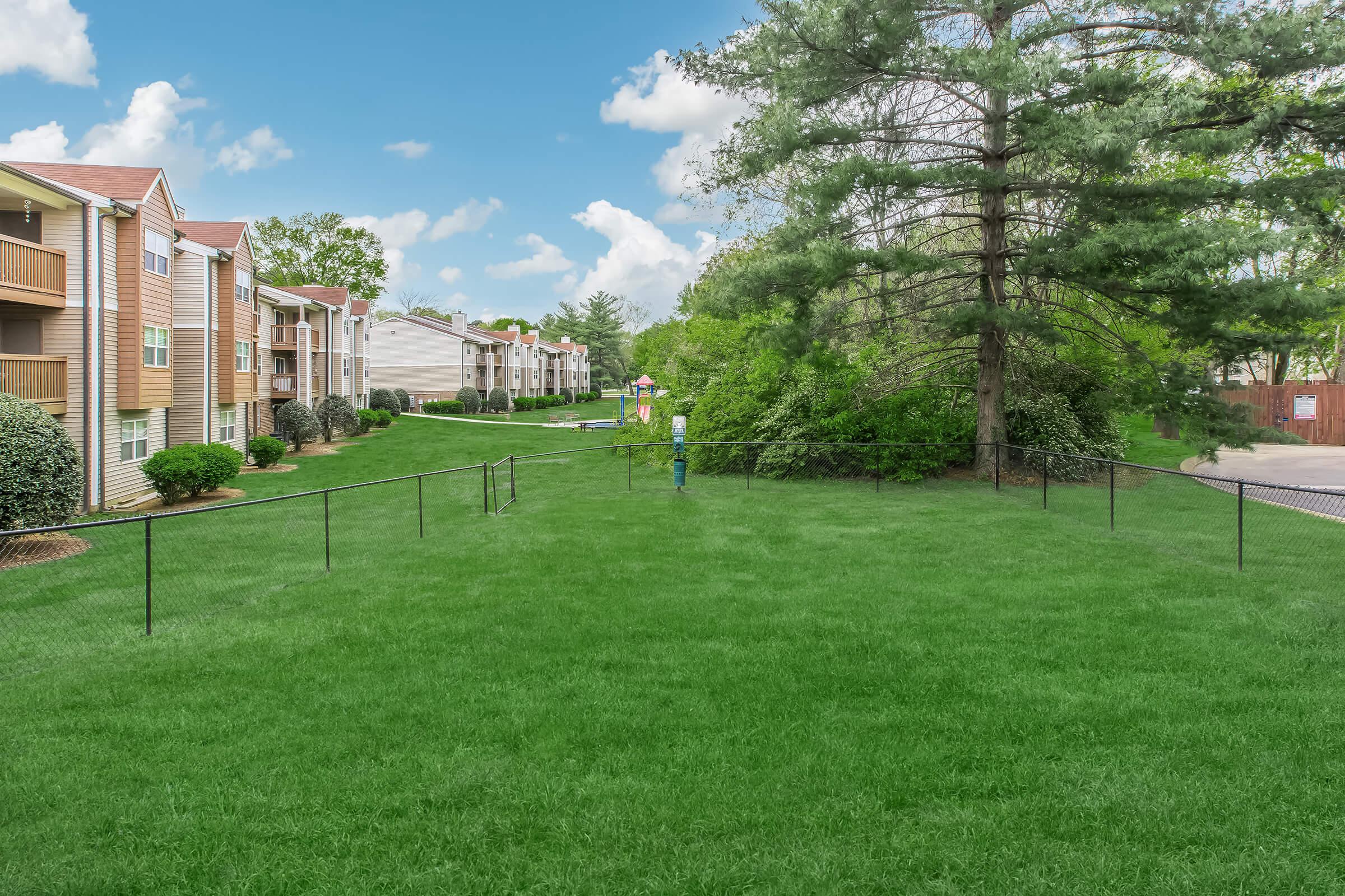 A grassy area surrounded by a black fence, with several apartment buildings in the background. Trees and shrubs are visible, and a person is walking along the path. The sky is clear with some clouds, creating a pleasant outdoor scene.