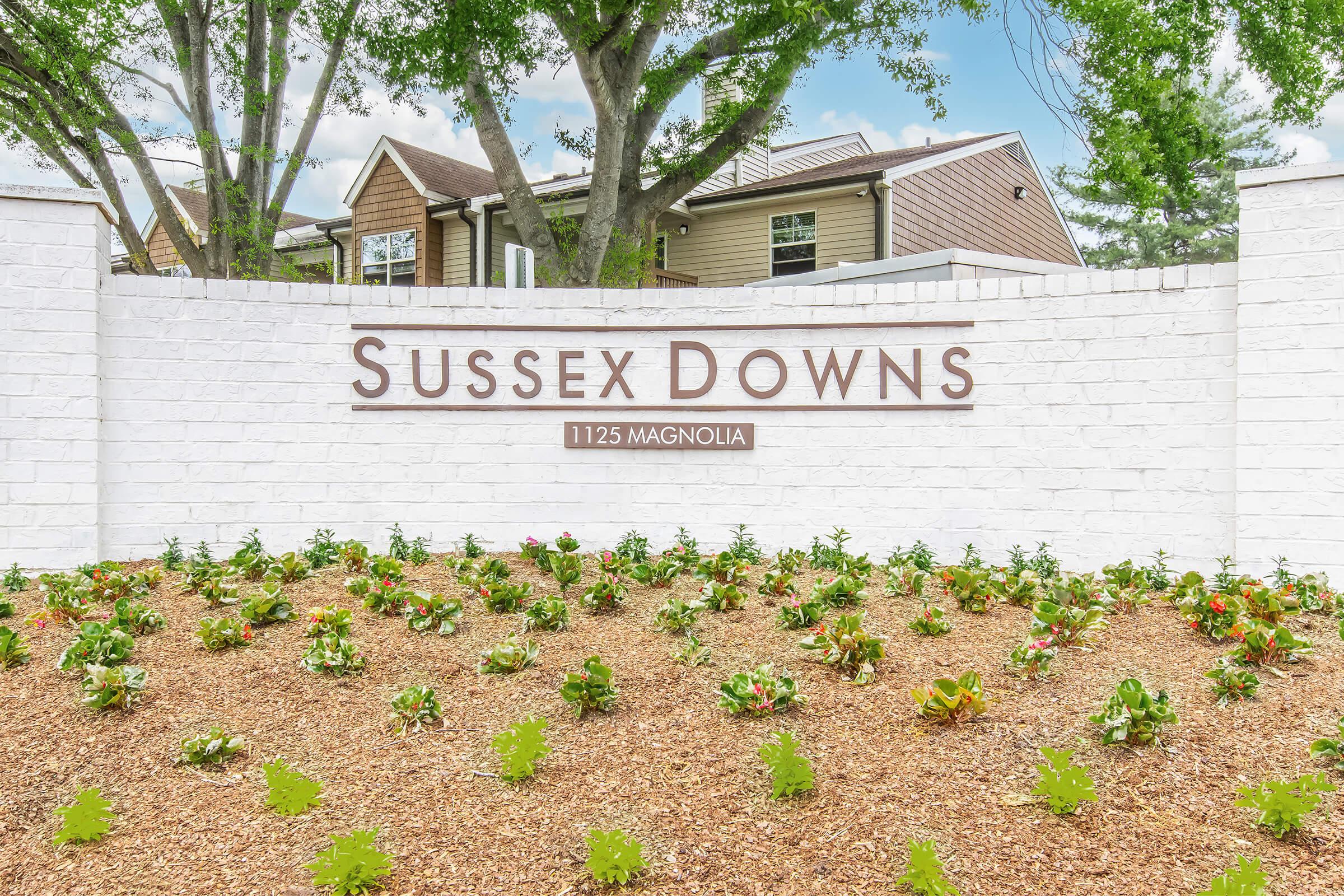 Sign featuring "Sussex Downs" with the address "1125 Magnolia," situated in front of a landscaped area with flowers and greenery. The background includes residential buildings and trees, indicating a welcoming community atmosphere.