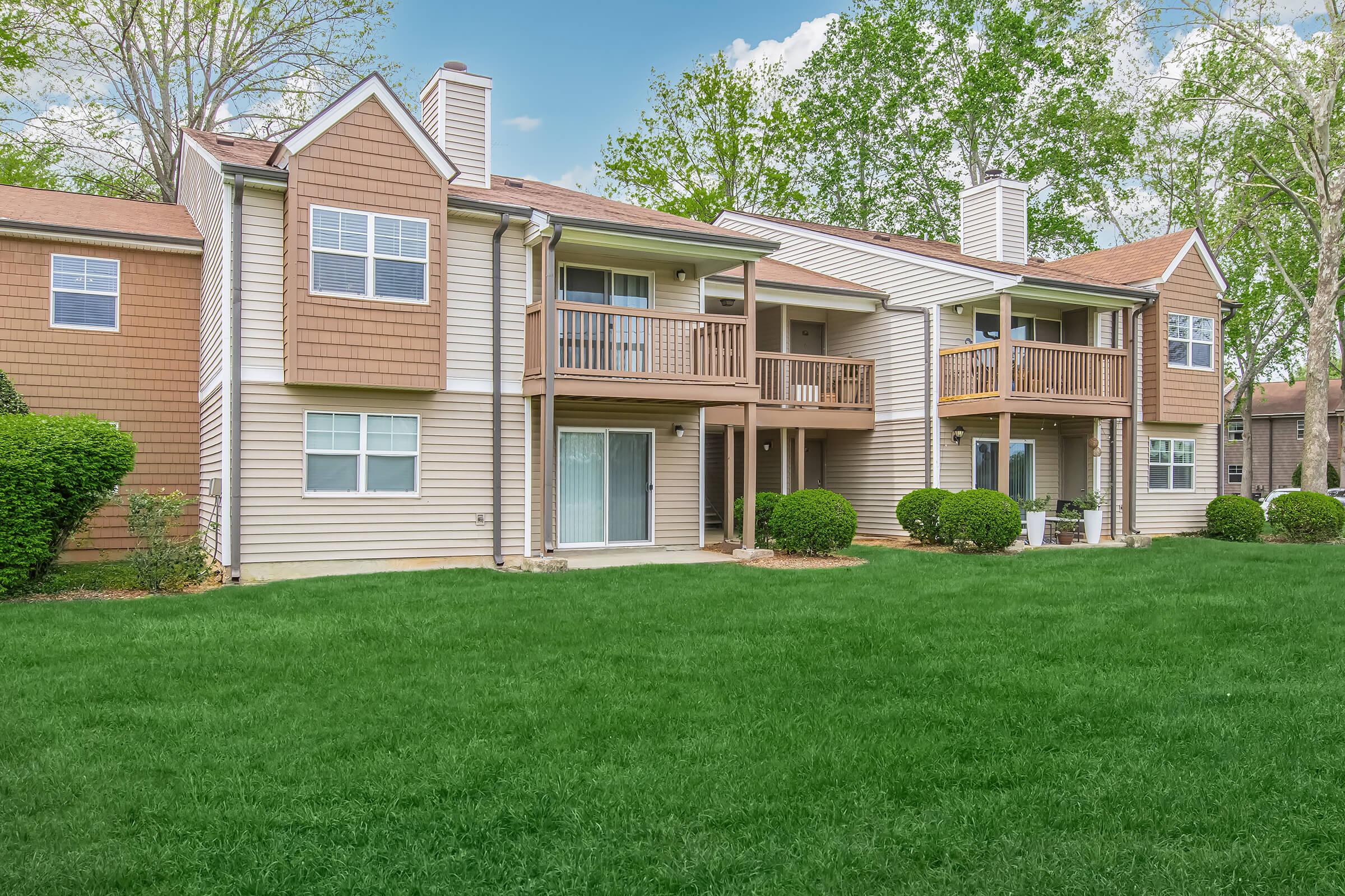 A welcoming view of a two-story residential building featuring balconies, with well-manicured green lawns and neatly trimmed bushes. The exterior showcases a combination of light and dark siding under a clear blue sky, suggesting a pleasant, sunny day.