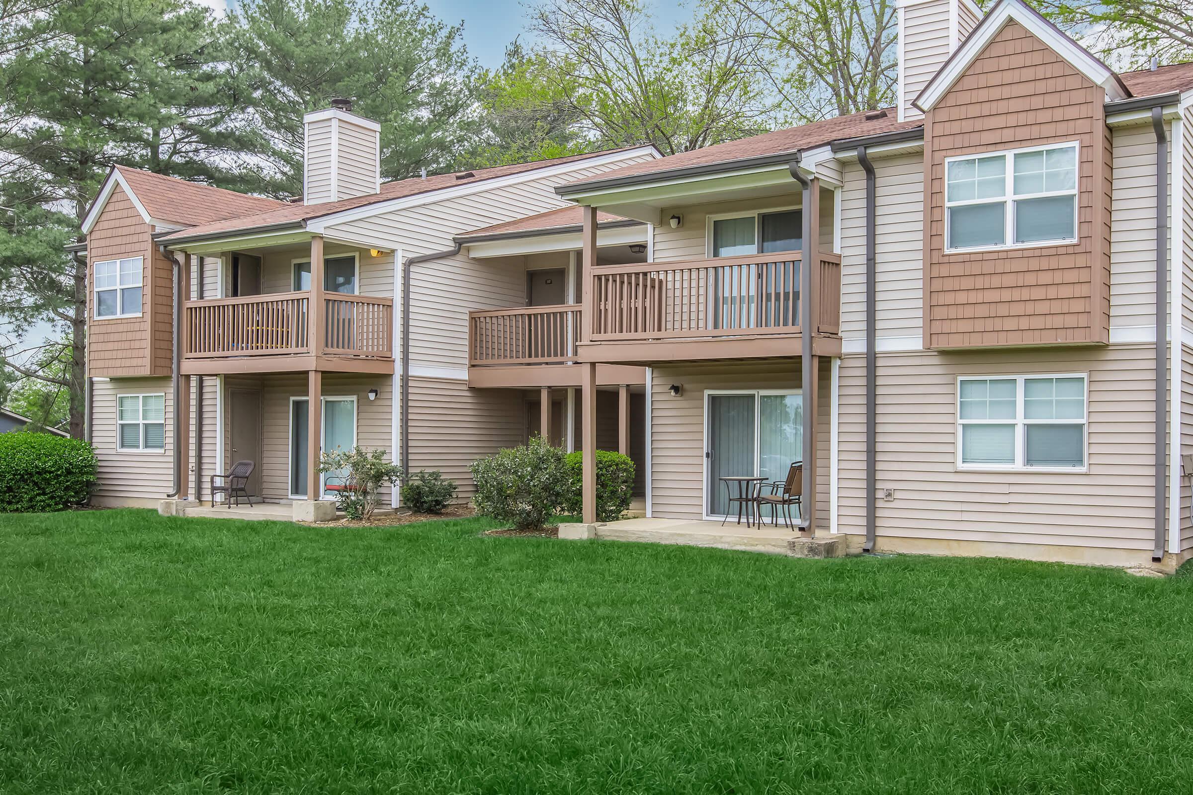 A two-story apartment building featuring balconies, with a well-maintained lawn in front. The siding is light brown with tan accents. Trees in the background provide greenery, and there are chairs on the balconies, indicating a pleasant outdoor space.