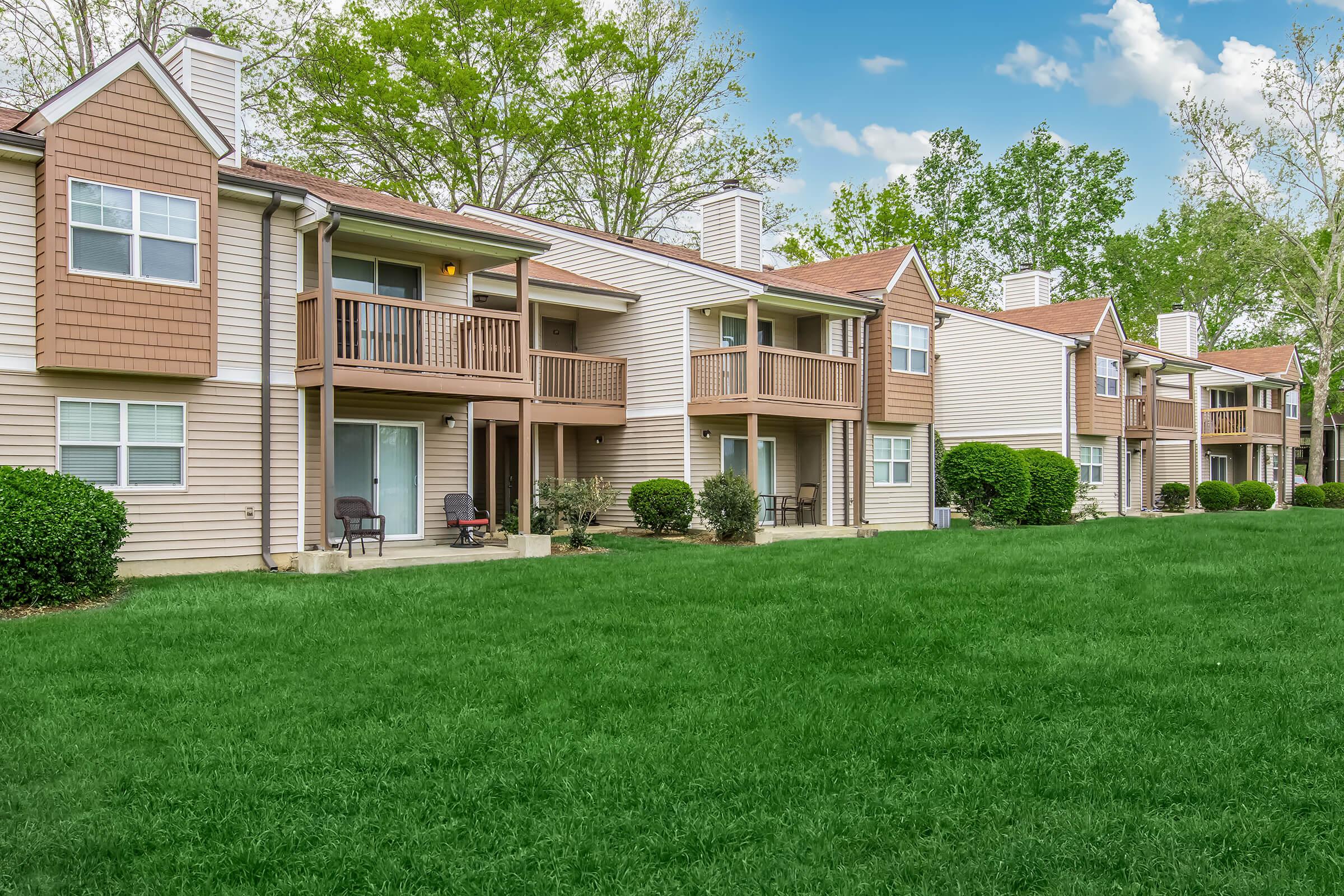 A row of multi-unit residential buildings with balconies, set in a green landscape. The buildings have light-colored siding and brown accents, surrounded by manicured lawns and trees under a sunny sky with a few clouds.