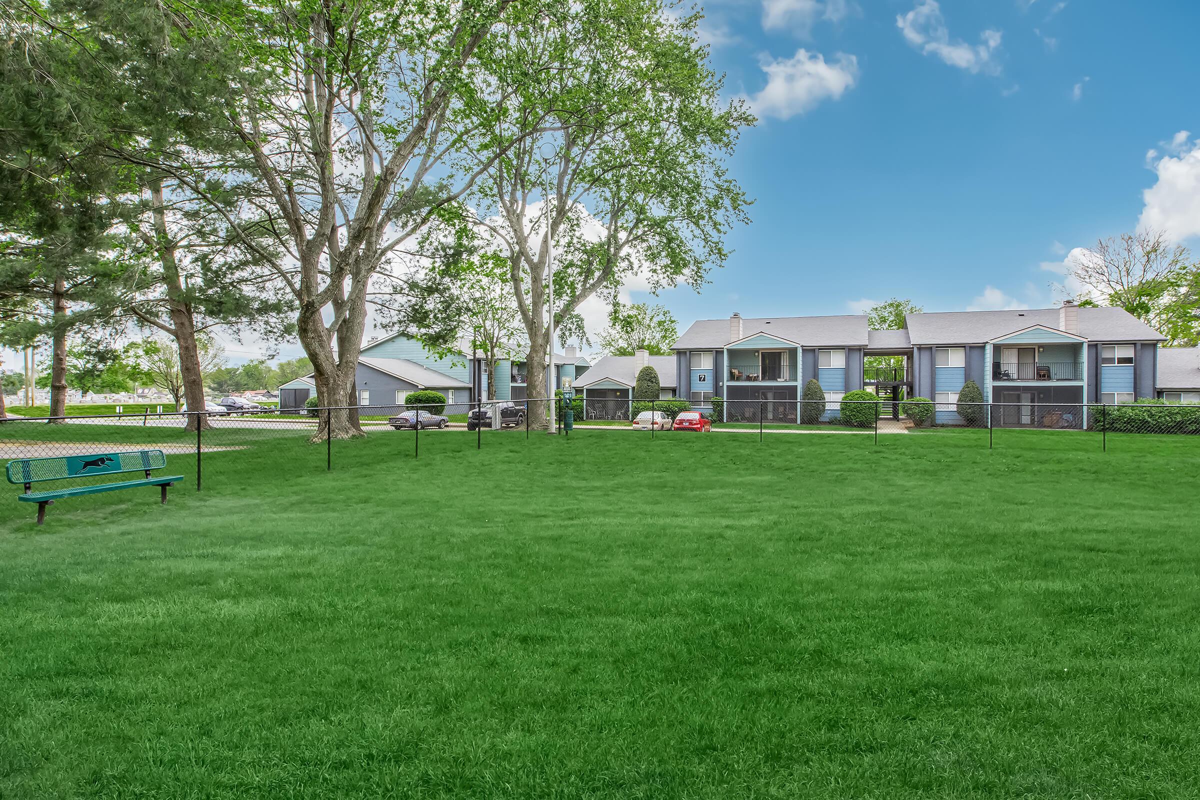 A lush green lawn is bordered by a fence, with several trees providing shade. In the background, a row of blue and gray residential buildings is visible, featuring balconies and parking spaces. The sky is bright blue with a few fluffy clouds. A bench is positioned on the grass looking toward the buildings.