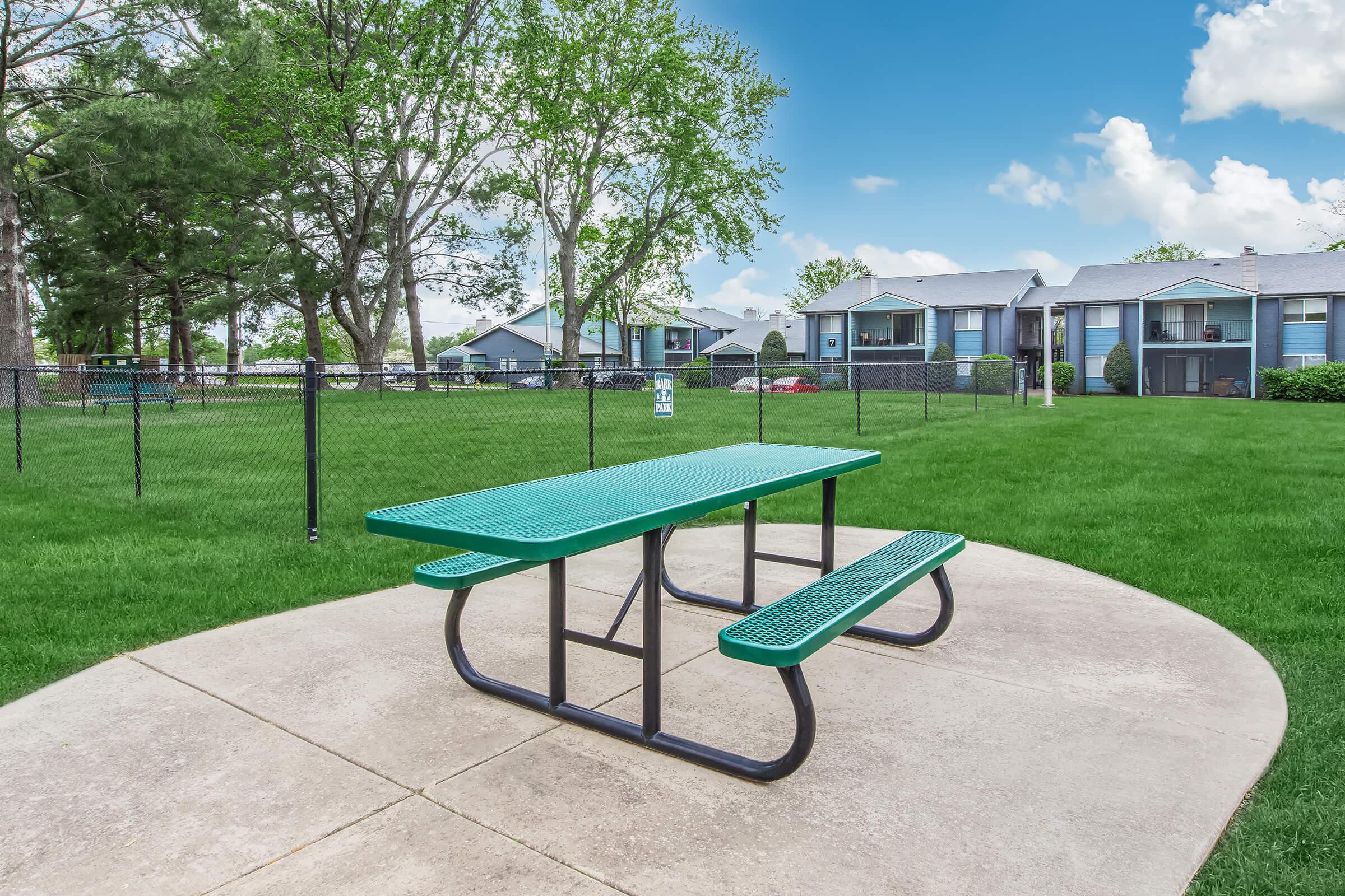 A green picnic table on a concrete pad in an open grassy area, surrounded by trees. In the background, there are several residential buildings, and a chain-link fence encloses the space, suggesting a community park or outdoor area. The sky is blue with some clouds.