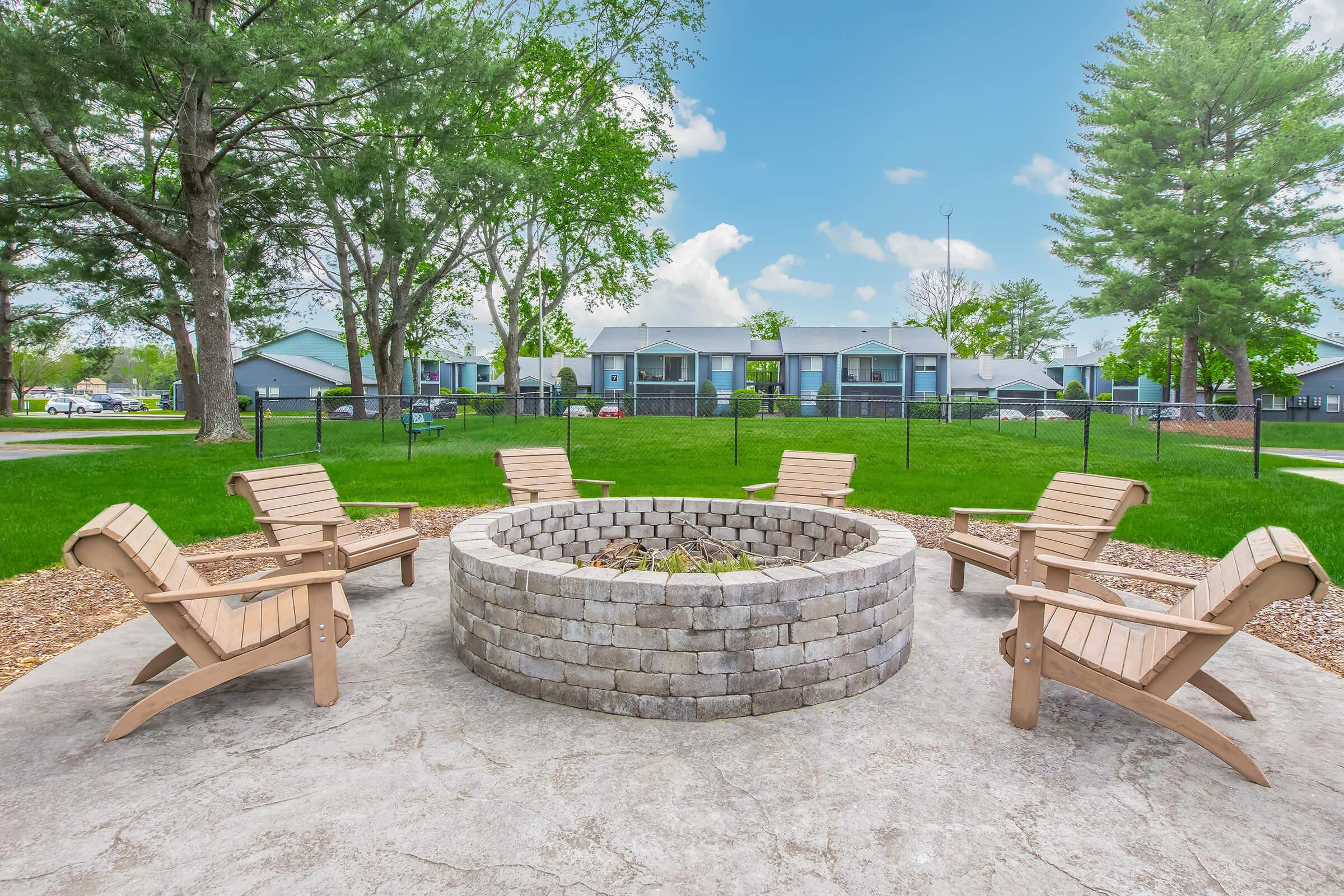 A circular stone fire pit surrounded by wooden chairs, set in a grassy area with trees and a blue sky. In the background, there are buildings, suggesting a residential or community space. The scene is inviting and designed for social gatherings.