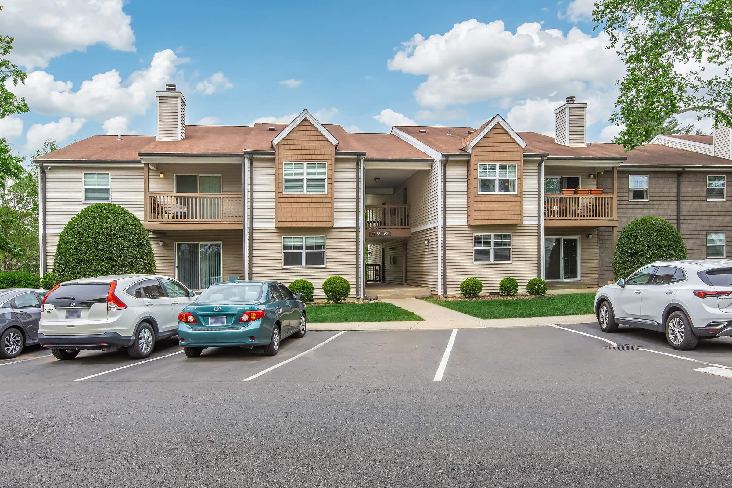 Two-story apartment building with a light brown exterior located in a residential area. The building features multiple balconies and well-maintained landscaping with bushes. In front, there is a parking lot with several parked cars, set against a backdrop of blue skies and scattered clouds.