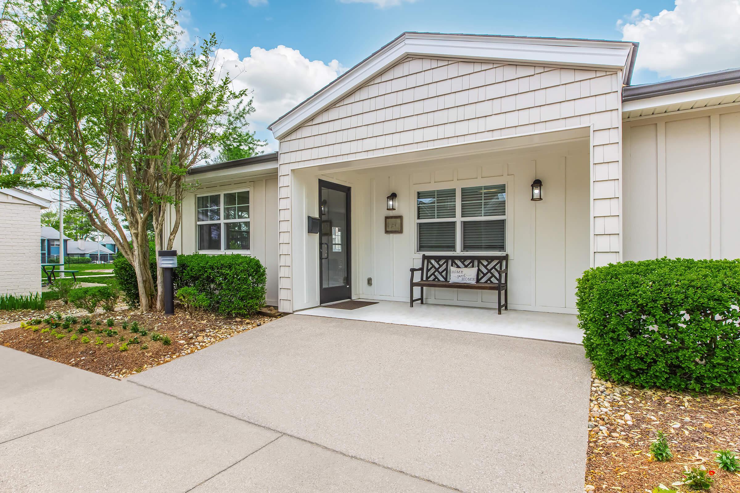 A modern, welcoming entrance to a light-colored house with a covered porch. There is a small bench and decorative plants on either side of the path leading to the door, framed by neatly trimmed bushes. The sky is bright with some clouds, enhancing the inviting atmosphere.