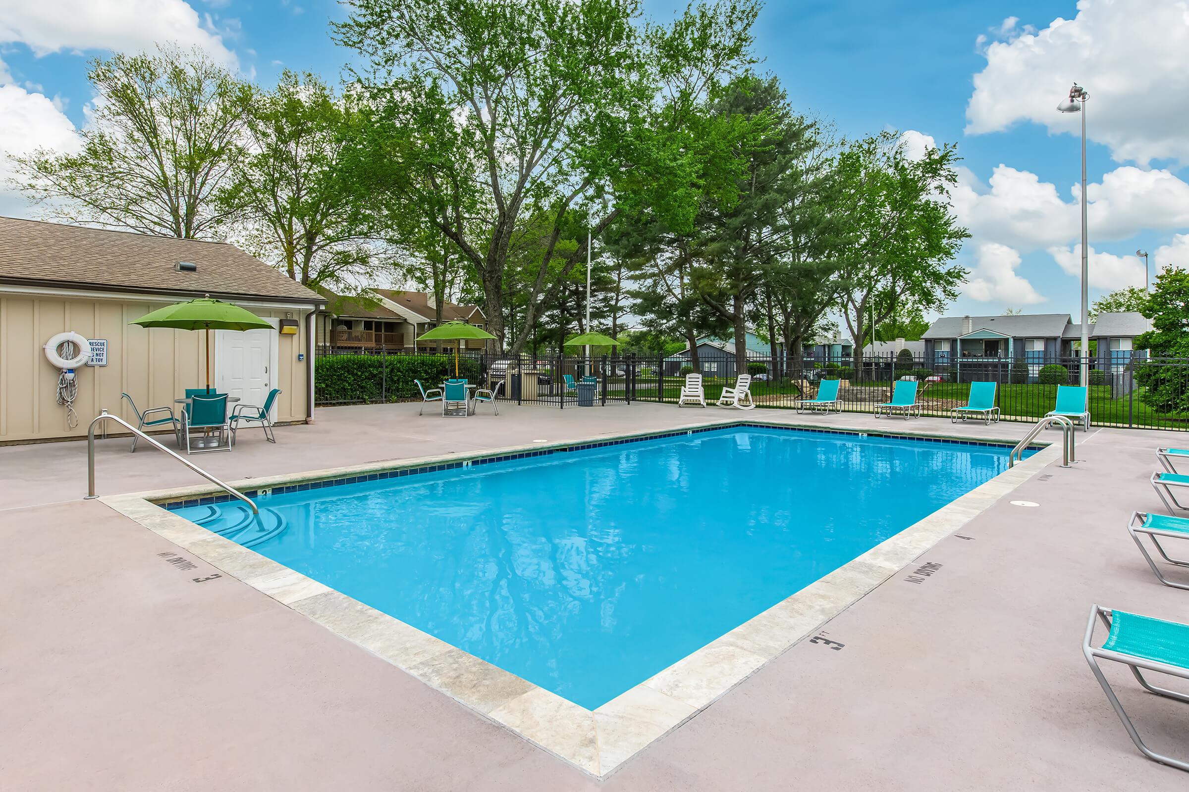 A clear blue swimming pool surrounded by green trees and sun loungers. There are umbrellas for shade nearby. A modest poolside area includes a building in the background, with a few chairs and a lifebuoy visible. The sky is bright with scattered clouds.