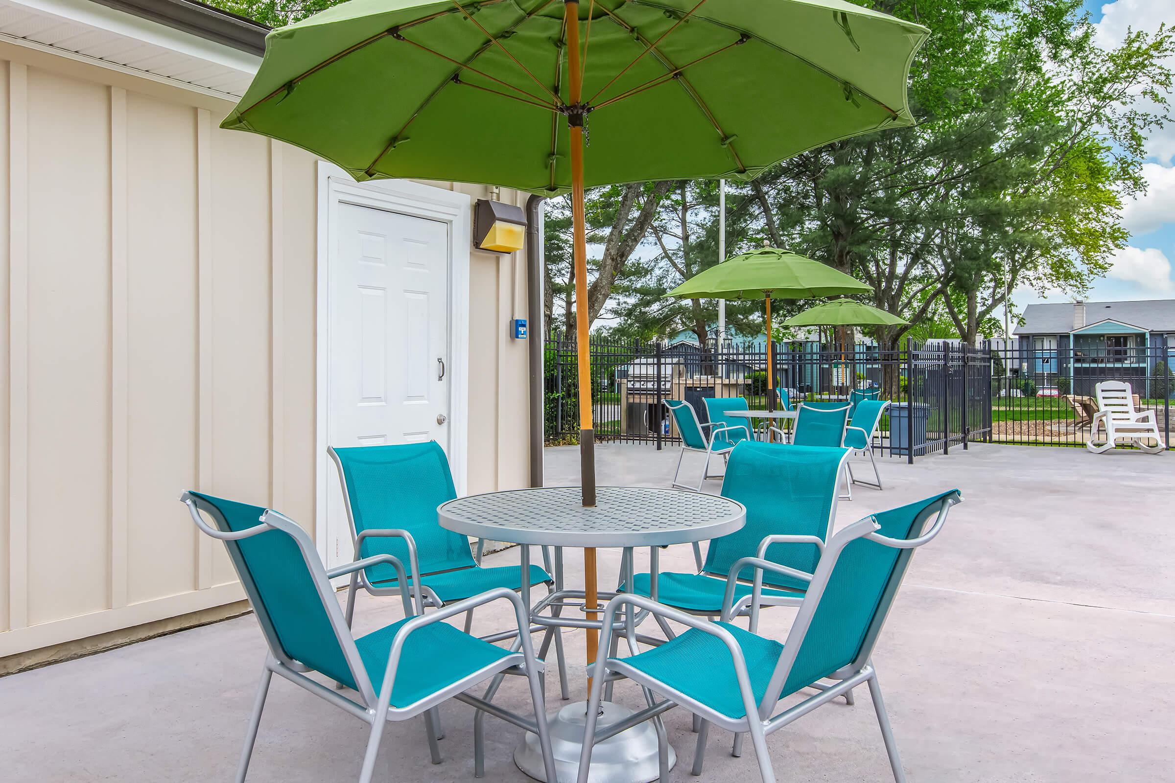 A patio area featuring a round table with a silver base surrounded by four teal chairs. Two green umbrellas provide shade, and there is a pool area visible in the background with lounge chairs and a fence. Trees and a blue sky add to the scene's inviting atmosphere.