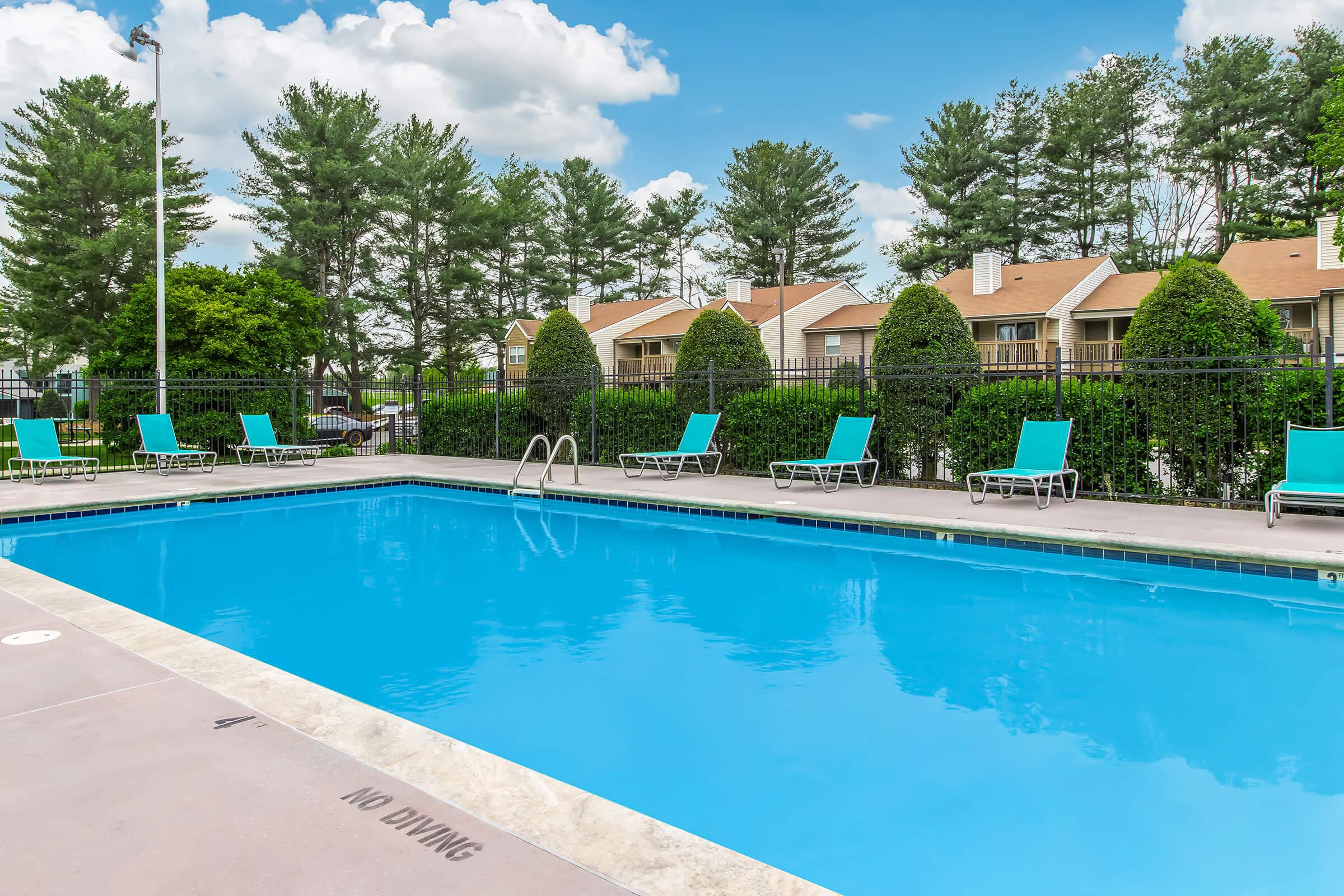 A clear blue swimming pool surrounded by green trees and well-maintained hedges. There are several lounge chairs with turquoise cushions positioned around the pool area. In the background, there are tan-colored residential buildings. The sky is partly cloudy.