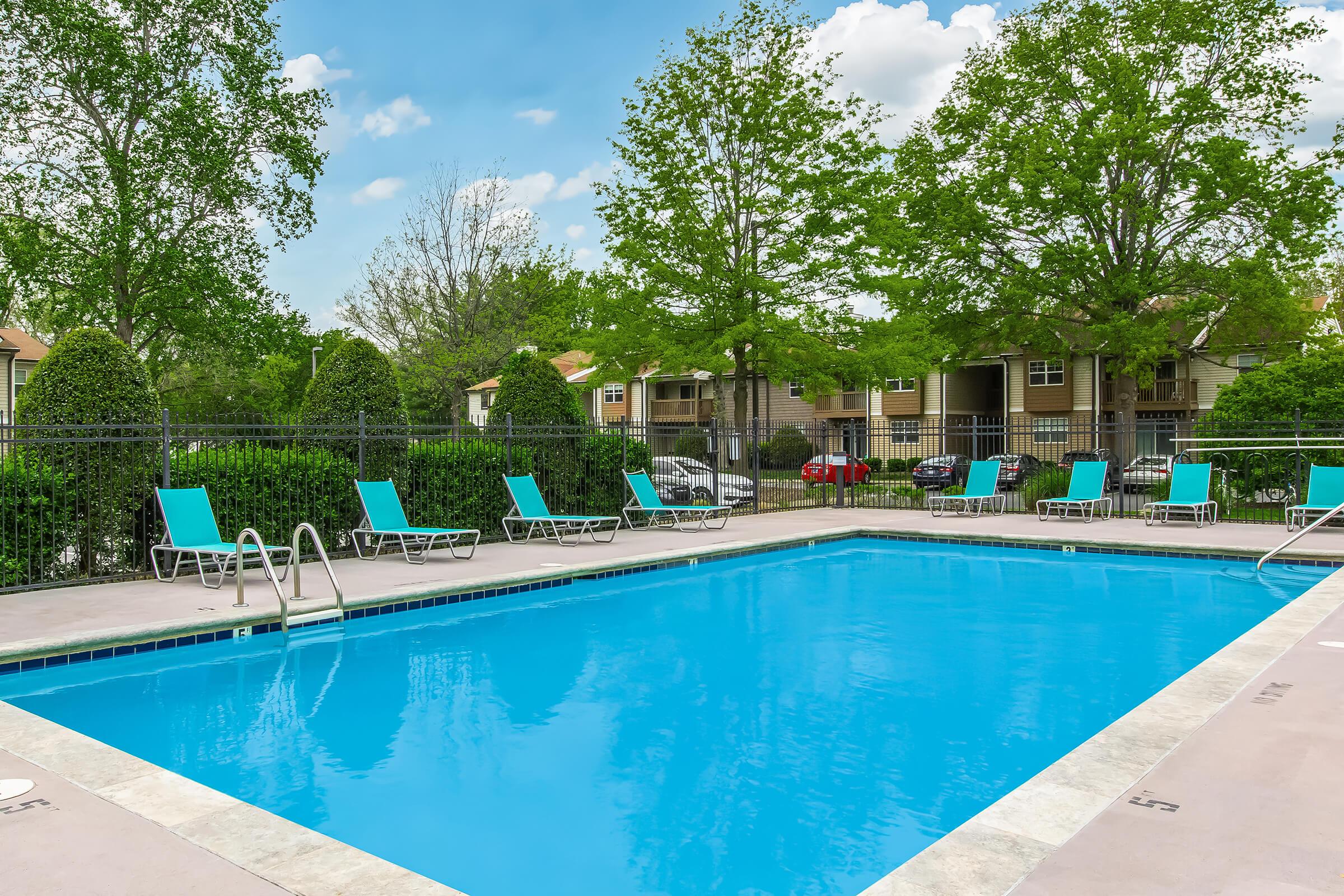A clear blue swimming pool surrounded by green trees and lounge chairs. In the background, there are residential buildings partially visible. The pool area is clean and well-maintained, creating a relaxing atmosphere on a sunny day.