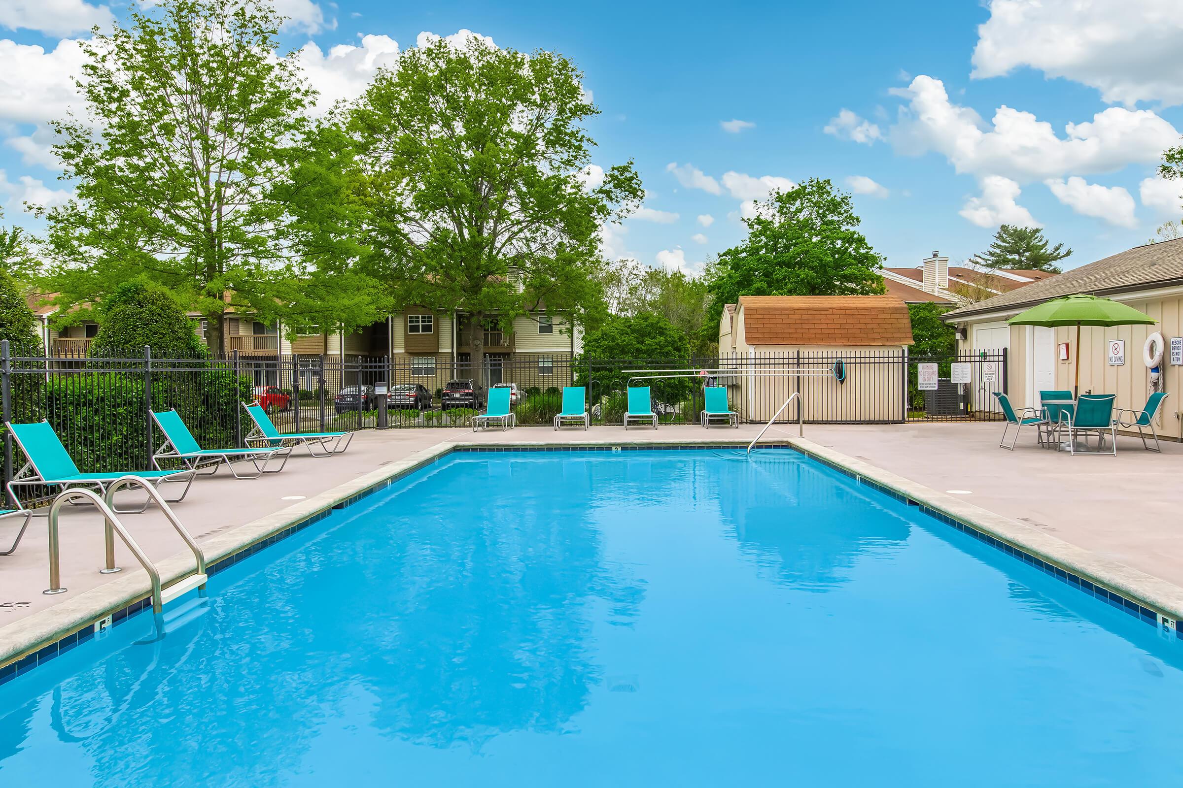 A vibrant outdoor pool with clear blue water, surrounded by green trees and lounge chairs. In the background, there are residential buildings and an umbrella-covered seating area. The sky is partly cloudy, adding to the relaxing atmosphere of the pool area.