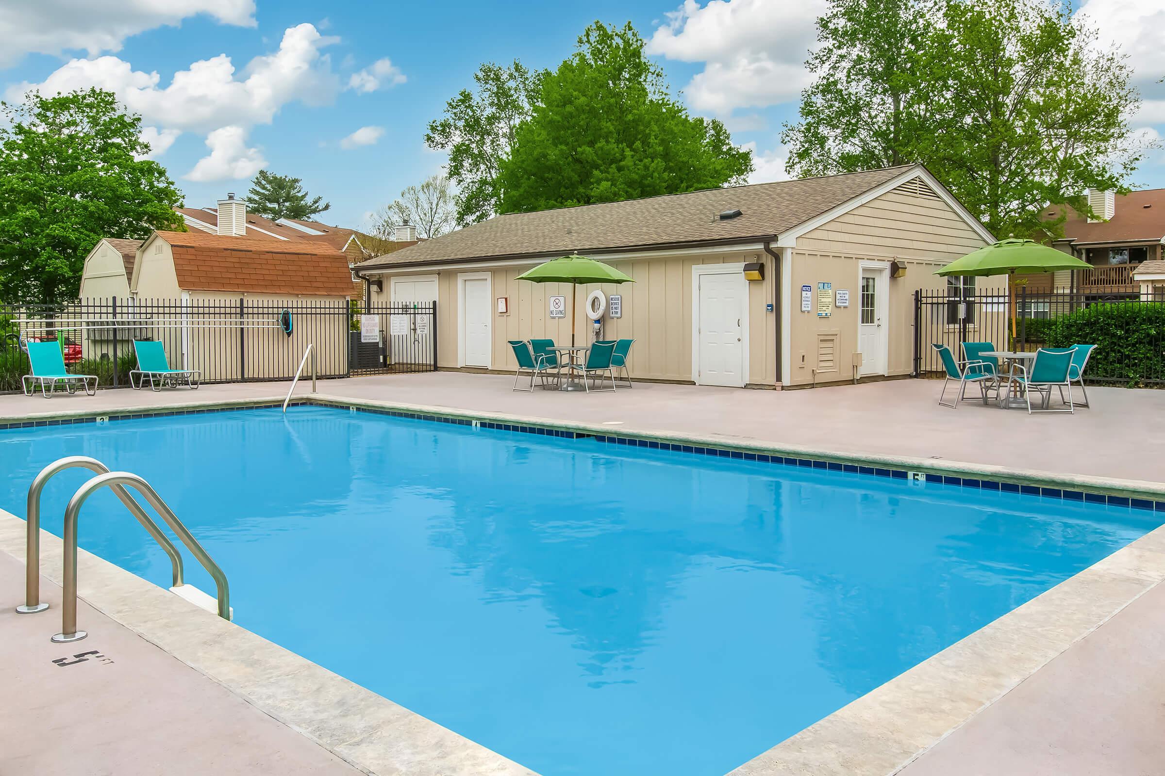 A clean outdoor swimming pool with a light blue surface, surrounded by a patio area featuring green umbrellas and lounge chairs. In the background, there is a small building and trees. The sky is partly cloudy, creating a pleasant atmosphere.