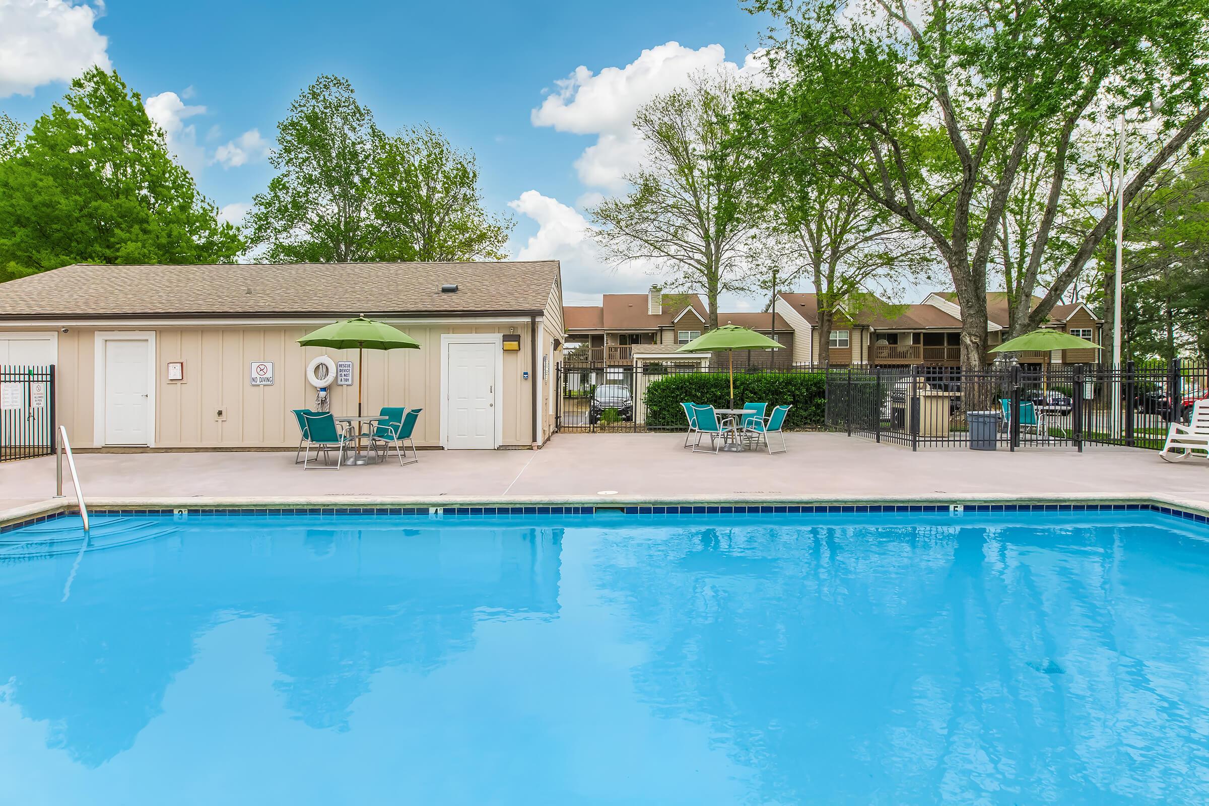 A clear blue swimming pool surrounded by a concrete patio. There are green umbrellas and chairs for seating. In the background, a building with multiple stories is visible, along with trees providing shade. The sky is bright with scattered clouds.