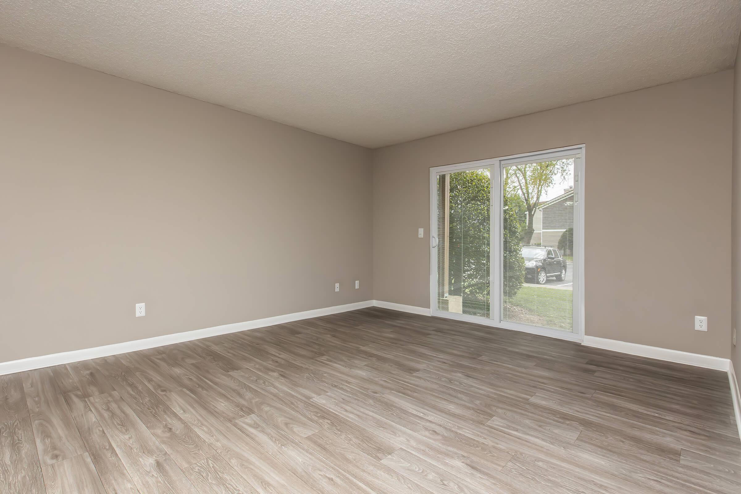 A spacious, empty room with light beige walls and modern vinyl flooring, featuring a sliding glass door that opens to an outdoor area with greenery. The room has a simple, uncluttered layout, creating an inviting and bright atmosphere.