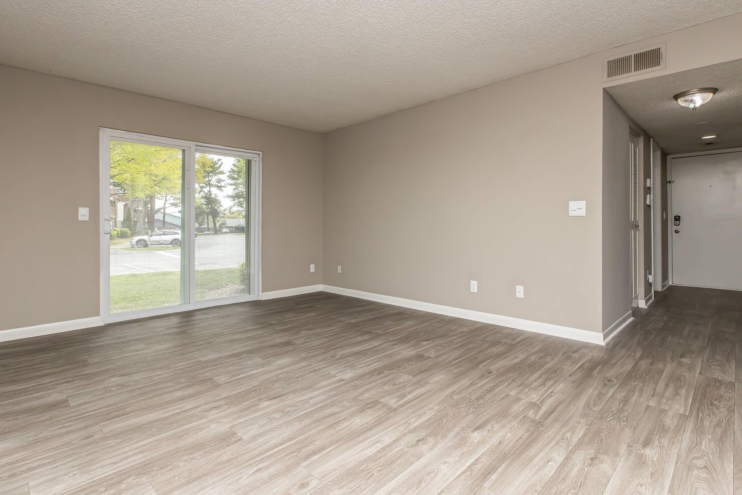 A spacious, empty living room with light gray walls and a light wood-style floor. A sliding glass door allows natural light and offers a view of the outdoors. The entrance is visible to the right, with a neutral color palette creating a bright, open atmosphere.