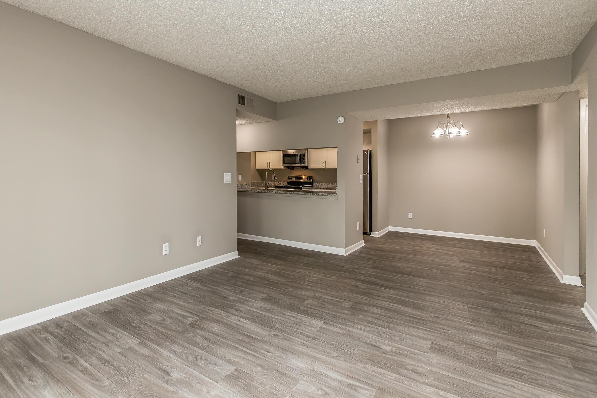 A spacious, modern living area featuring light grey walls and hardwood-style flooring. In the background, a kitchen is visible with stainless steel appliances. A dining area with a chandelier is seen on the right, creating a warm and inviting atmosphere.