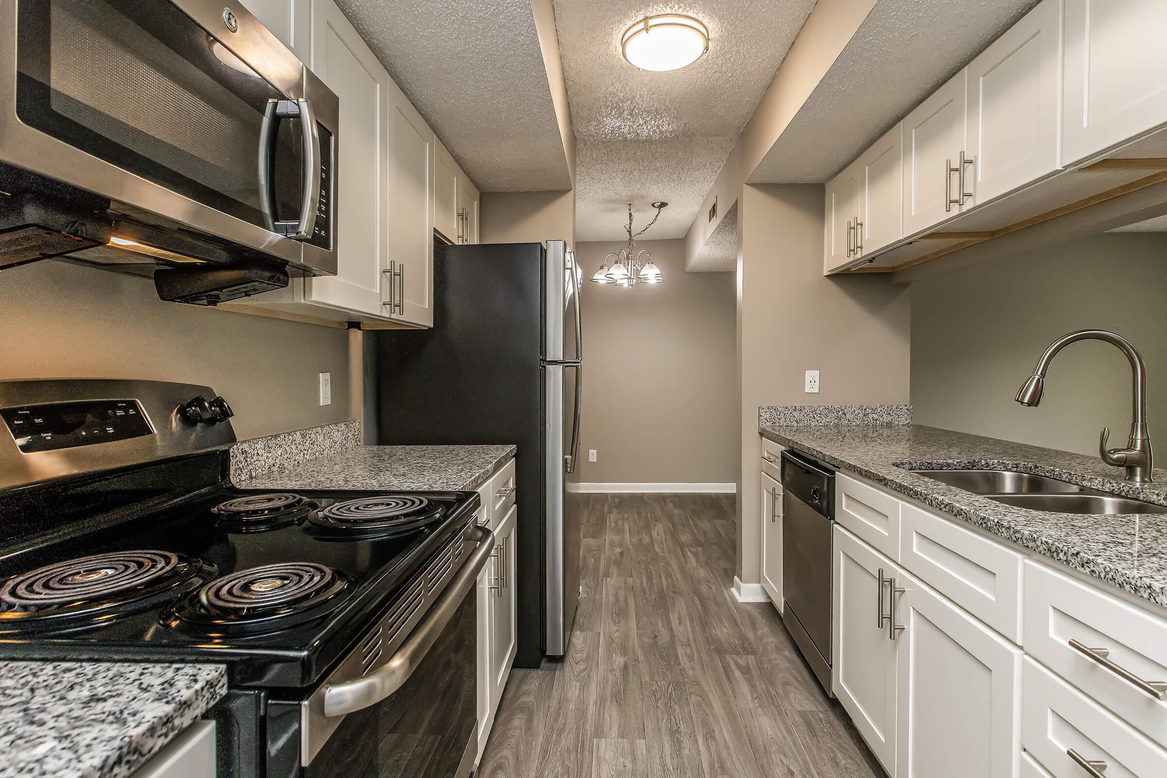 A modern kitchen featuring stainless steel appliances, including a microwave and refrigerator. The countertops are finished in grey granite, and the cabinets are light-colored. The flooring is a wood-like laminate, adding warmth to the space. A light fixture is visible, enhancing the ambiance.