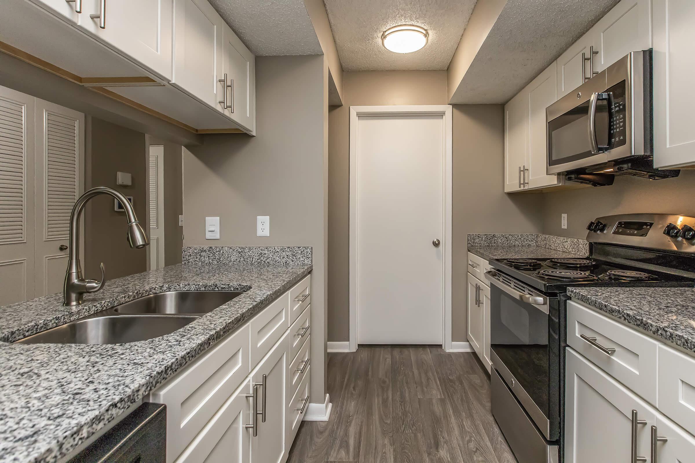 A modern kitchen featuring white cabinetry, a granite countertop, stainless steel appliances including a microwave and stove, and a sink with a faucet. The flooring is a wood-like laminate, and there is a door leading to another room at the end of the kitchen space.