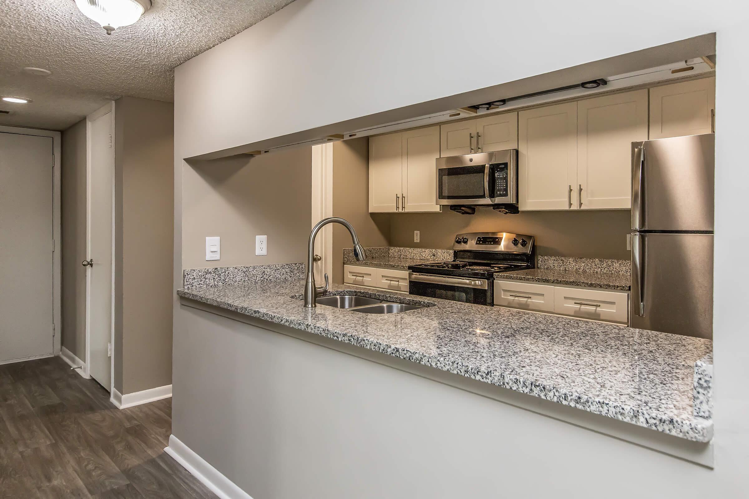 Modern kitchen featuring granite countertops, stainless steel appliances, and an open design. The kitchen includes a double sink and is connected to a cozy living area. Light-colored cabinets and a neutral wall color enhance the spacious feel. Bright overhead lighting complements the overall aesthetic.