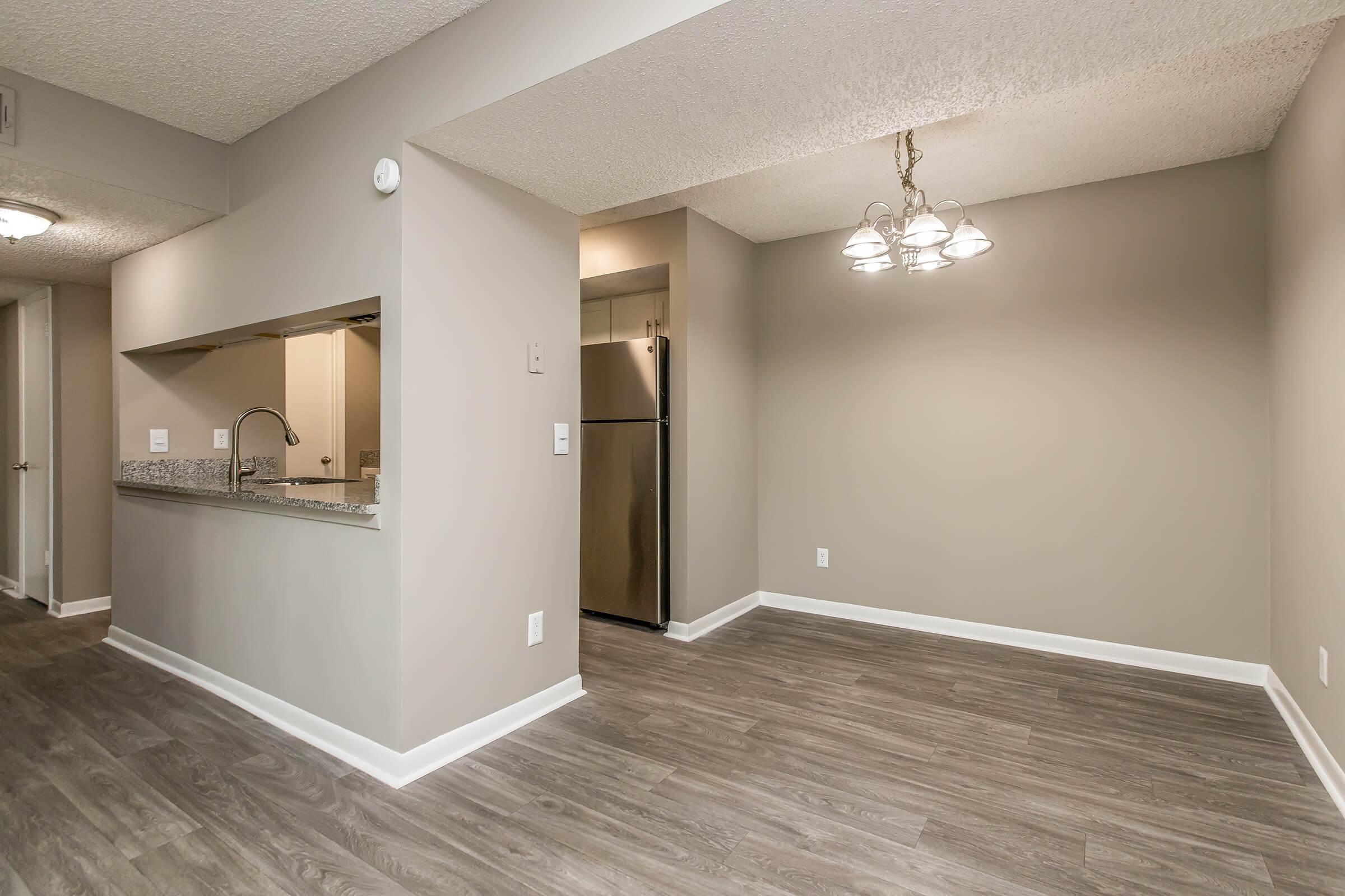 A modern living space featuring a light gray color scheme. The room includes a small kitchen area with a breakfast bar, a stainless steel refrigerator, and a dining area illuminated by a chandelier. The flooring is a wood-like laminate, enhancing the contemporary atmosphere.