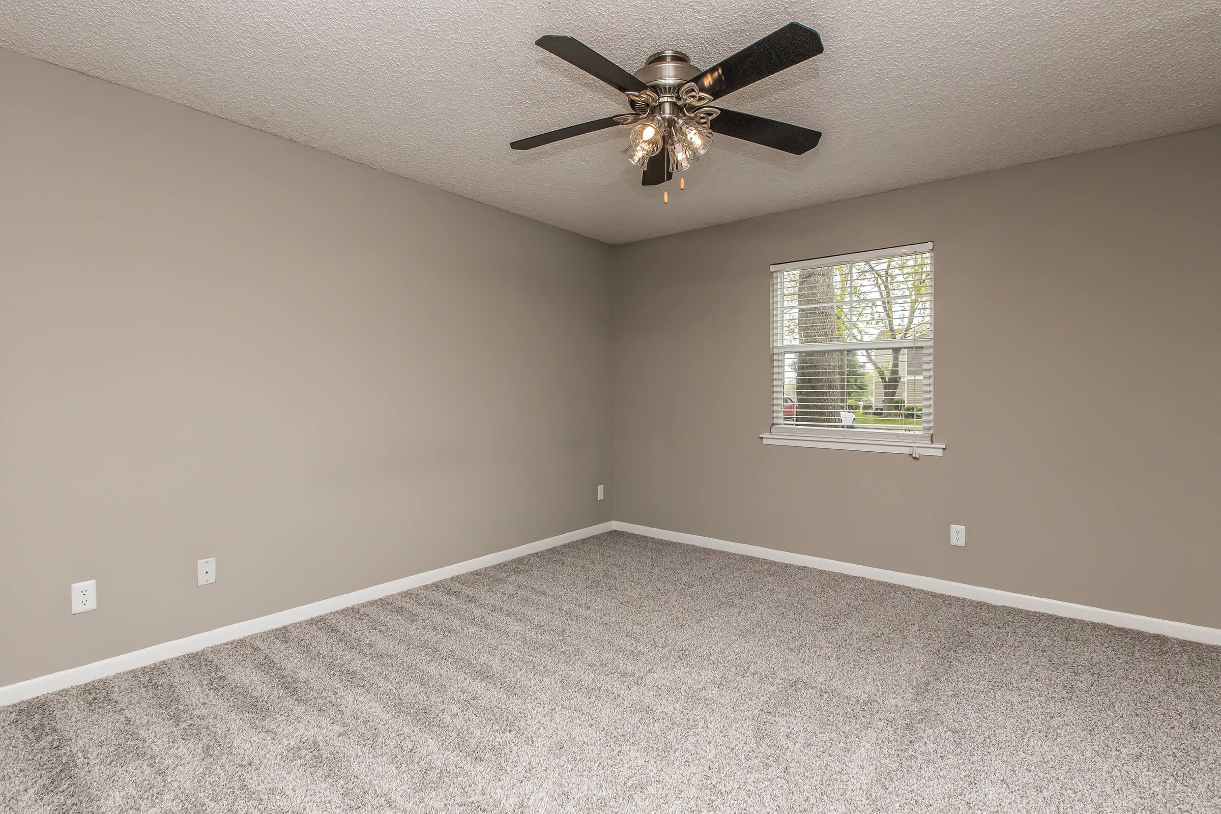 A spacious, empty room with light gray walls and a plush carpet. The ceiling features a ceiling fan with five blades. A window with white blinds allows natural light to enter, and there are two electrical outlets on the walls. The room is unfurnished, providing a blank canvas for decoration.